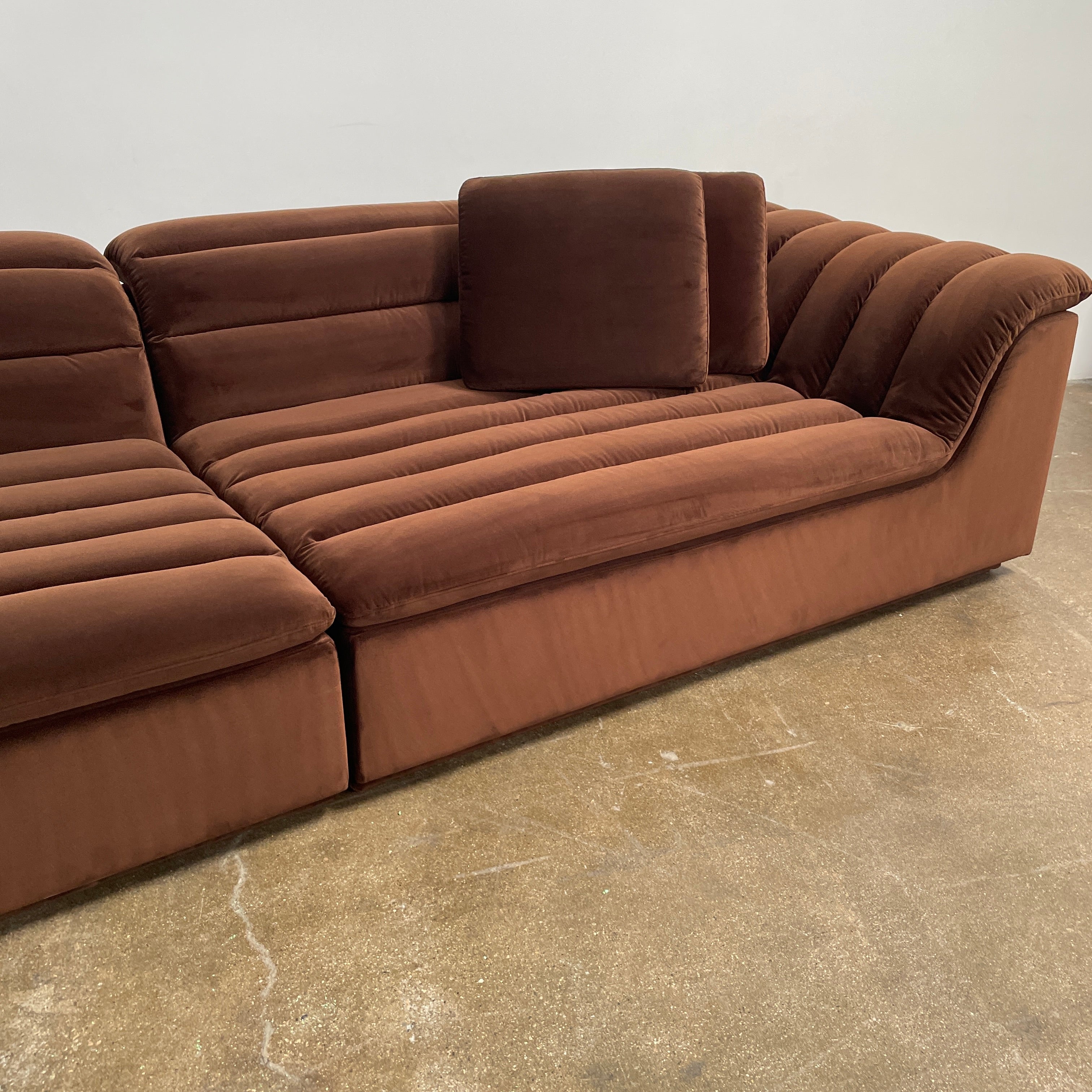 The Ellison Studios Float Sofa is a dark brown sectional from Ellison Studios with velvet upholstery, low profile, channel tufting, and two large cushions for extra comfort.
