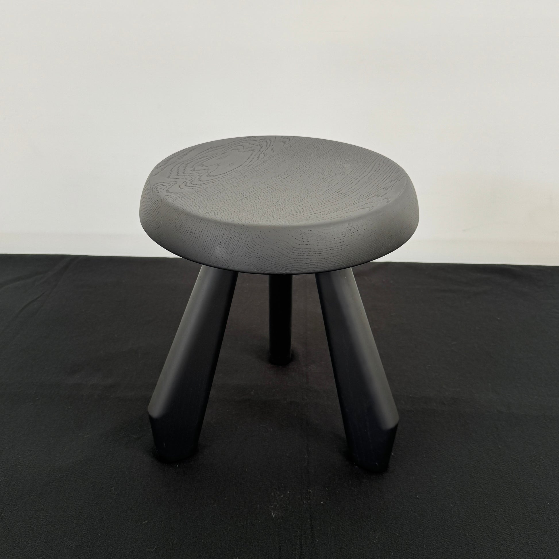 The Cassina 523 Tabouret Meribel by Cassina is a small, round, three-legged solid wood stool with a gray seat and black legs, shown against a plain white background.