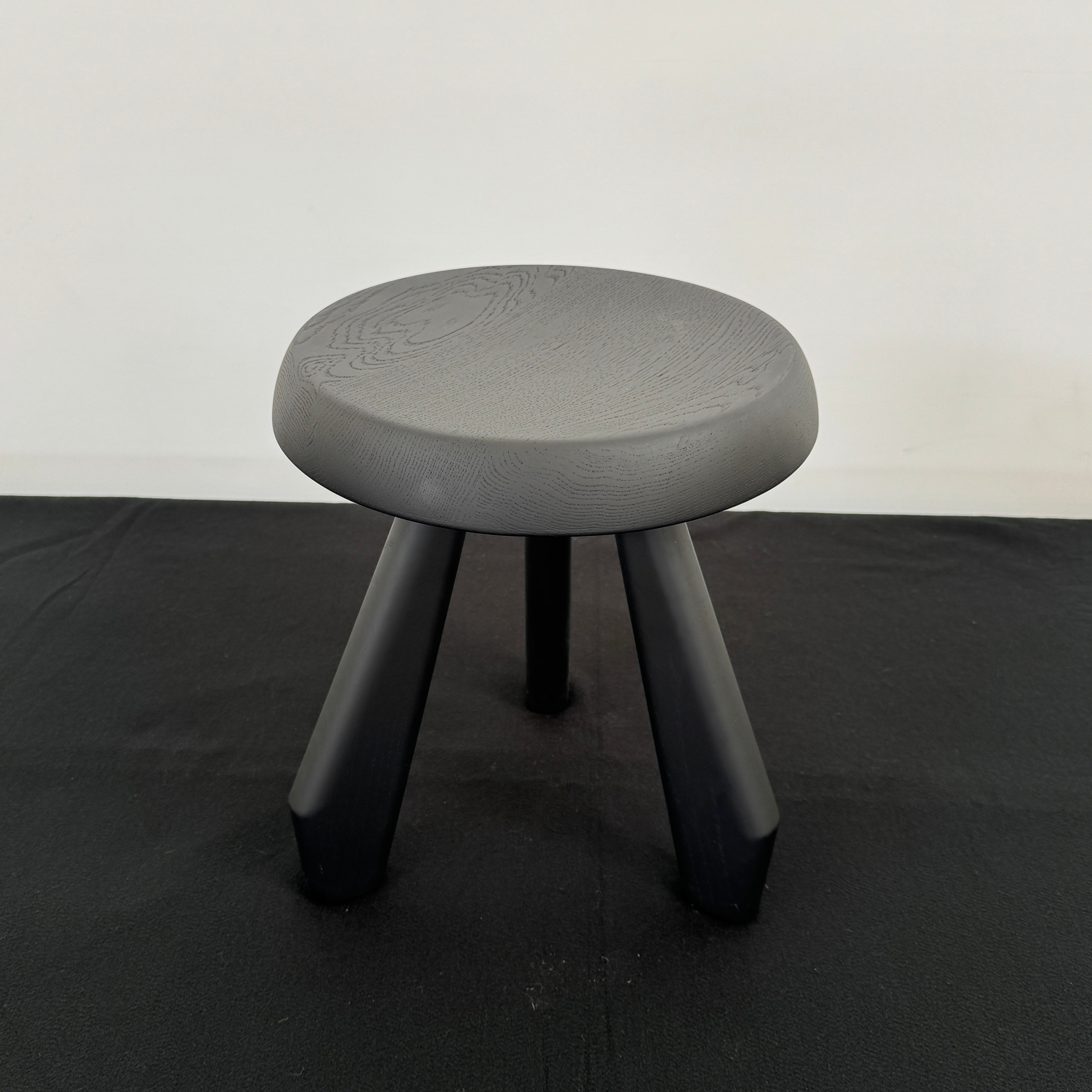 The Cassina 523 Tabouret Meribel by Cassina is a small, round, three-legged solid wood stool with a gray seat and black legs, shown against a plain white background.