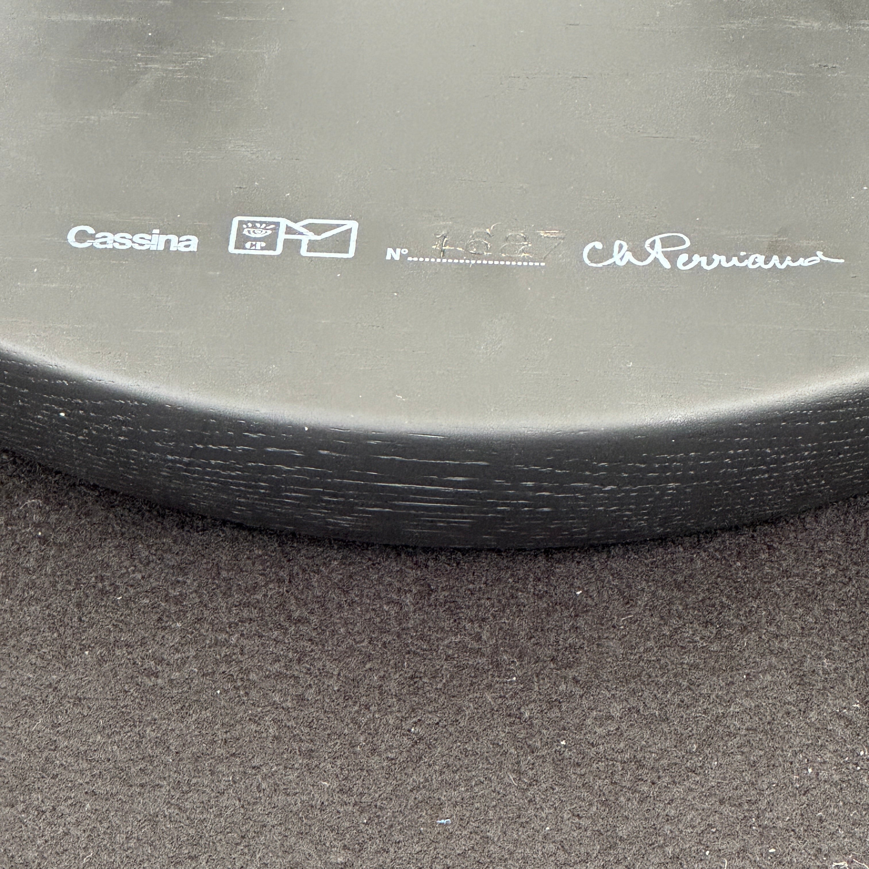 The Cassina 523 Tabouret Meribel by Cassina is a small, round, three-legged solid wood stool with a gray seat and black legs, shown against a plain white background.