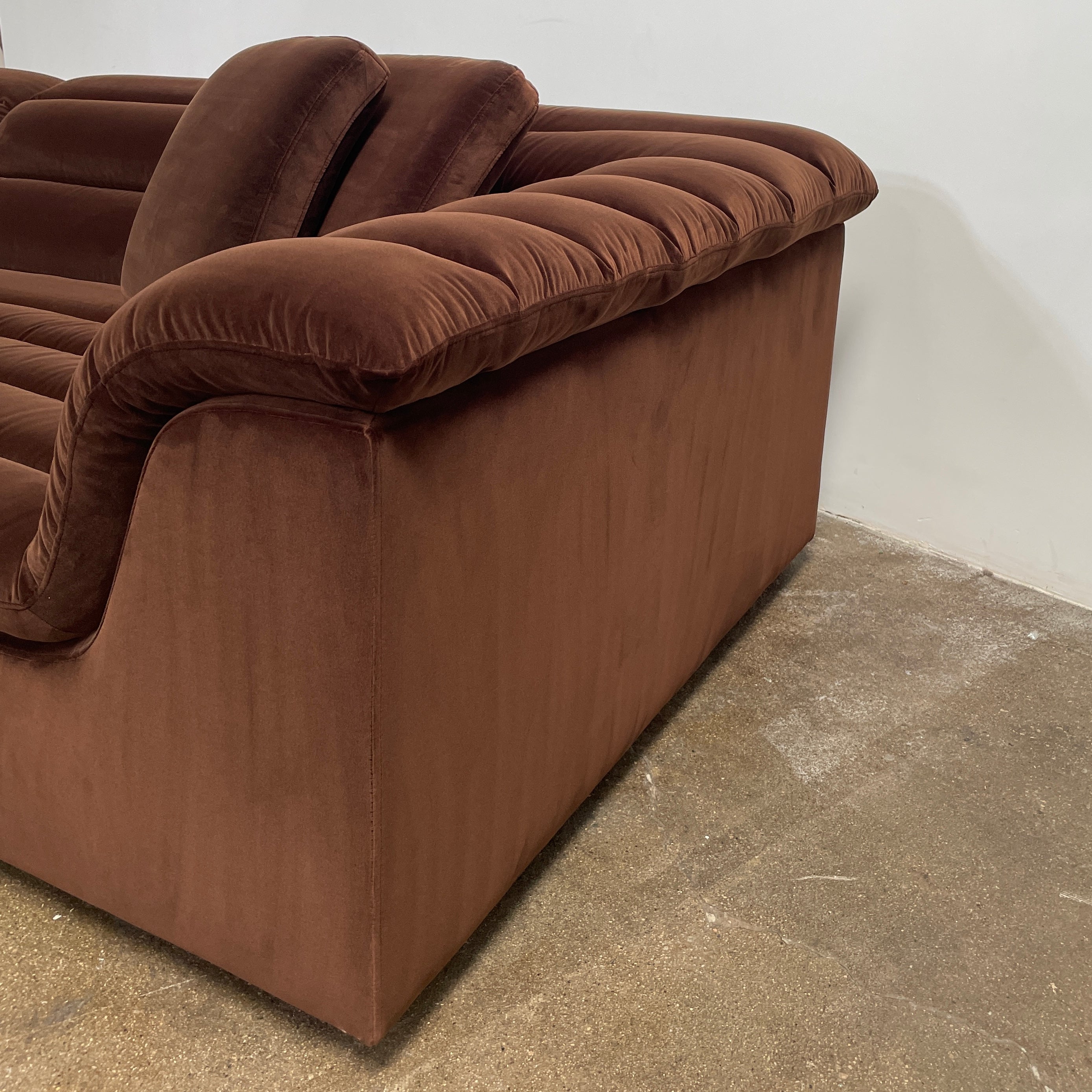 The Ellison Studios Float Sofa is a dark brown sectional from Ellison Studios with velvet upholstery, low profile, channel tufting, and two large cushions for extra comfort.