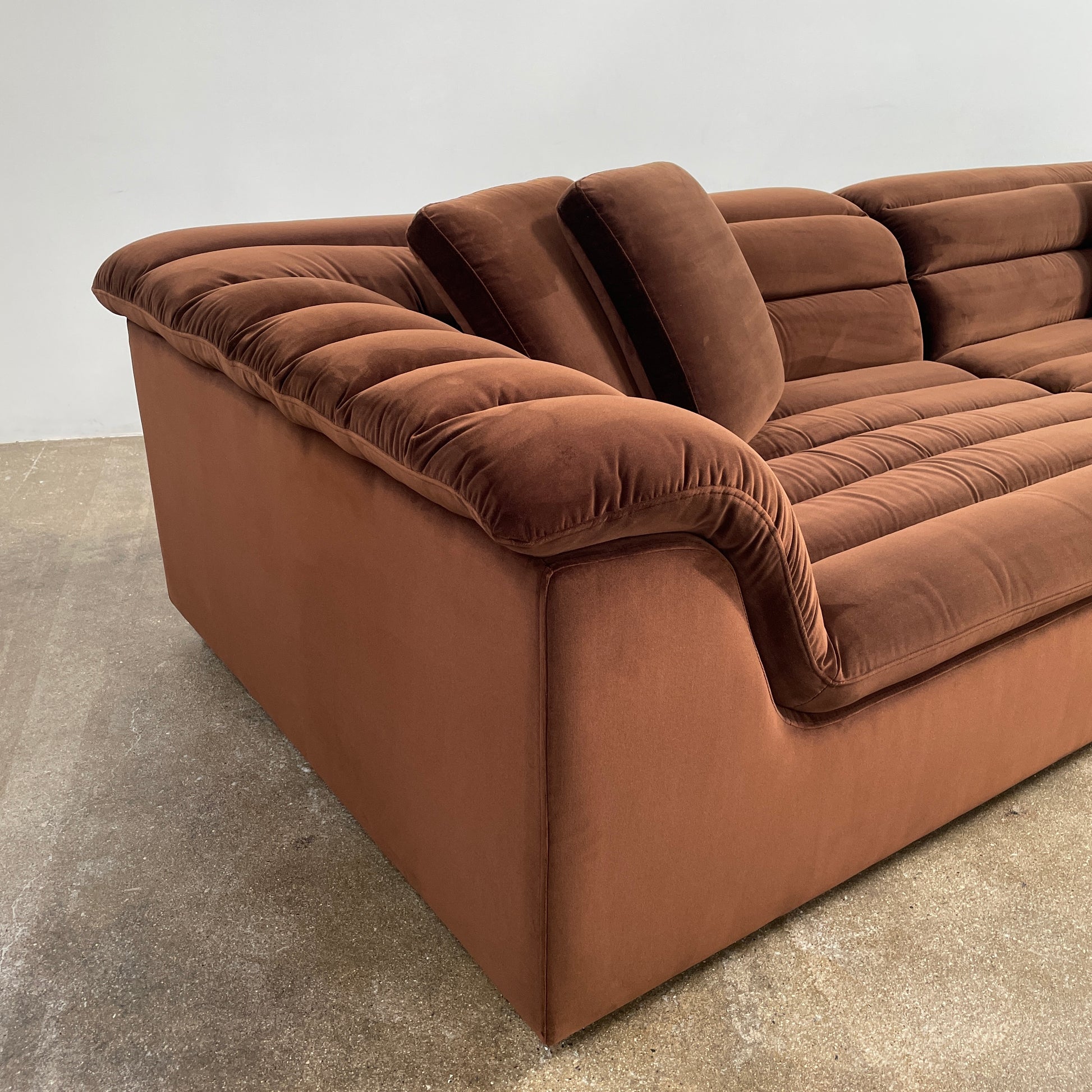 The Ellison Studios Float Sofa is a dark brown sectional from Ellison Studios with velvet upholstery, low profile, channel tufting, and two large cushions for extra comfort.