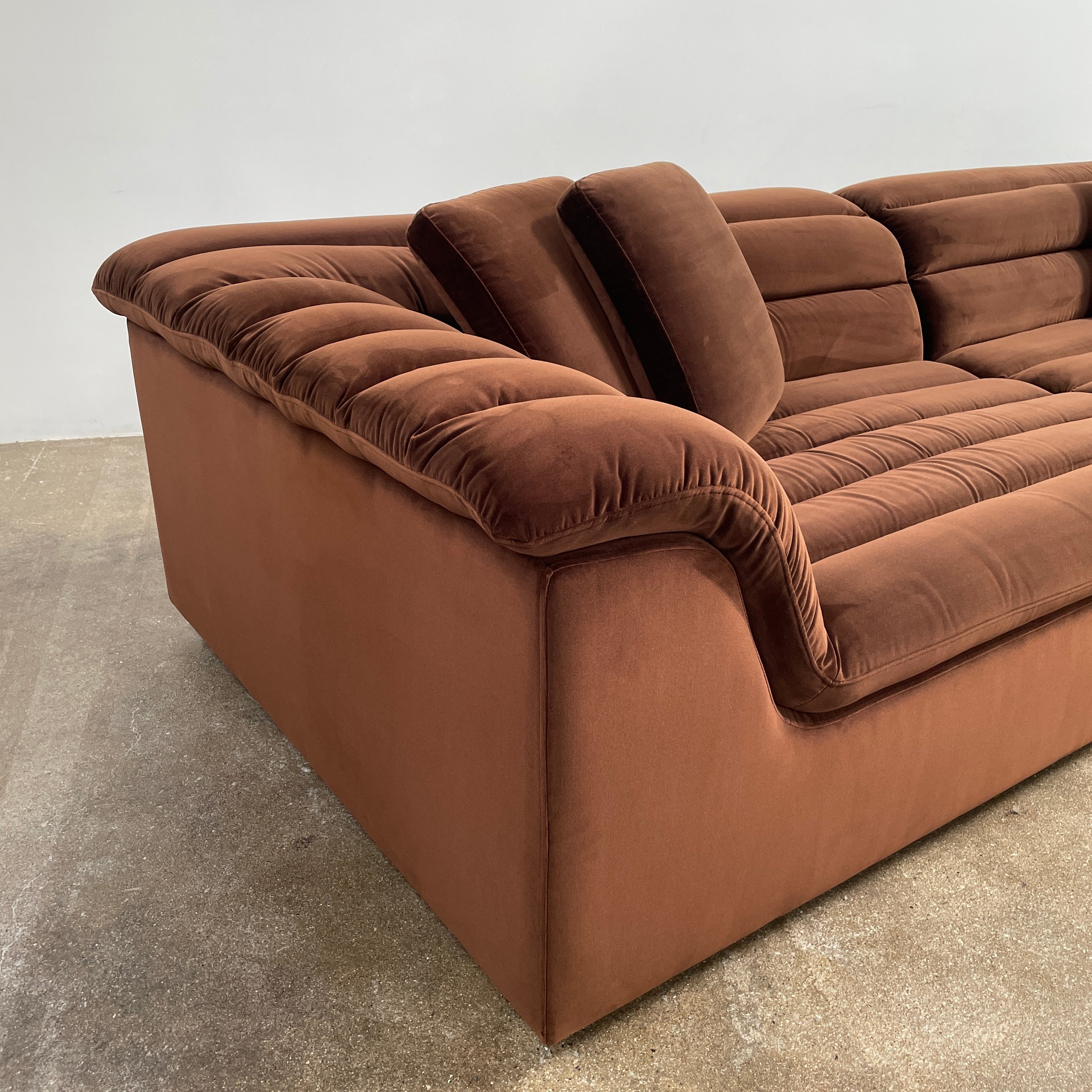 The Ellison Studios Float Sofa is a dark brown sectional from Ellison Studios with velvet upholstery, low profile, channel tufting, and two large cushions for extra comfort.