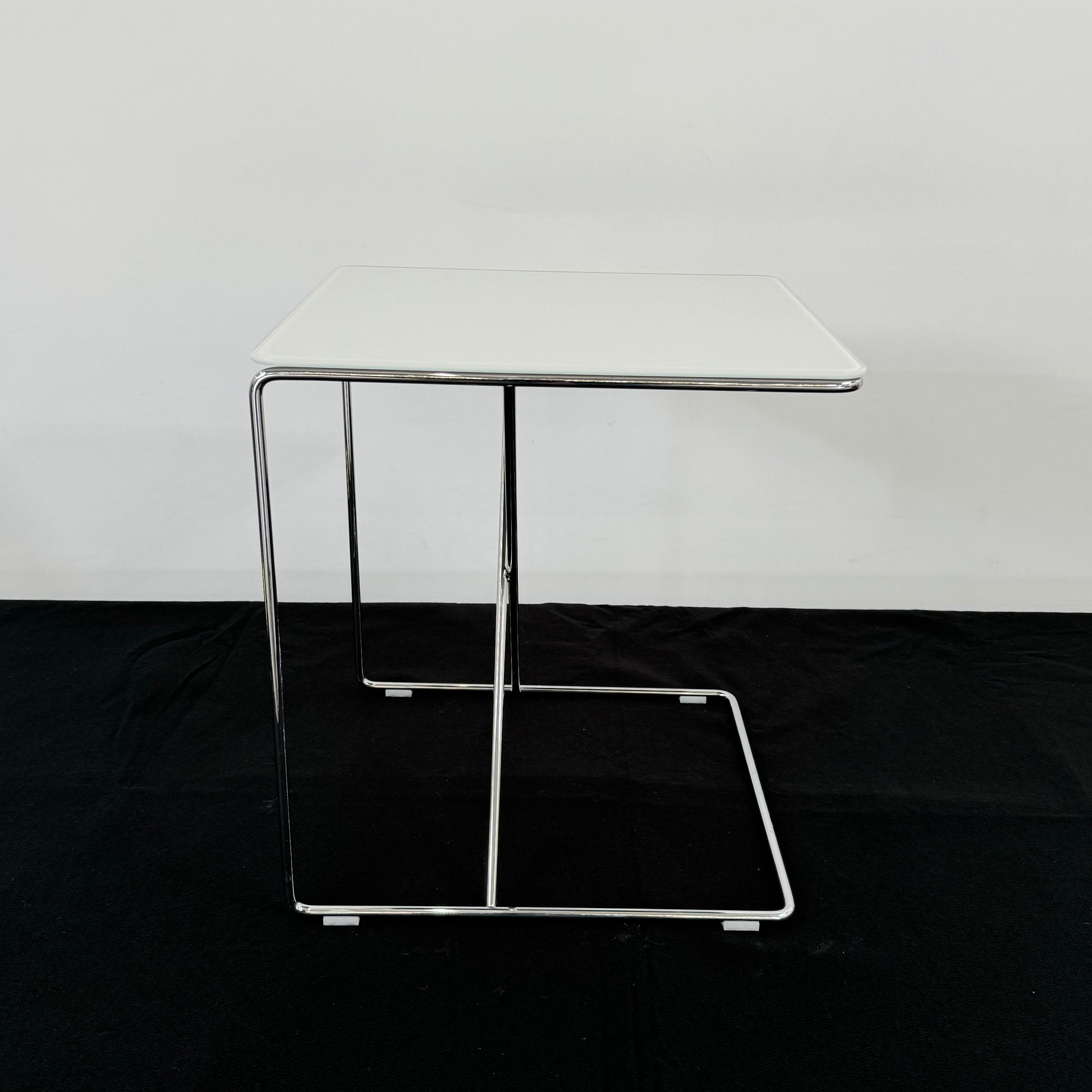 The B&B Italia JJ Cantilever Accent Table by B&B Italia features a white rectangular top and minimalist chrome frame, ideal for modern spaces. Shown against a plain white wall and black floor.