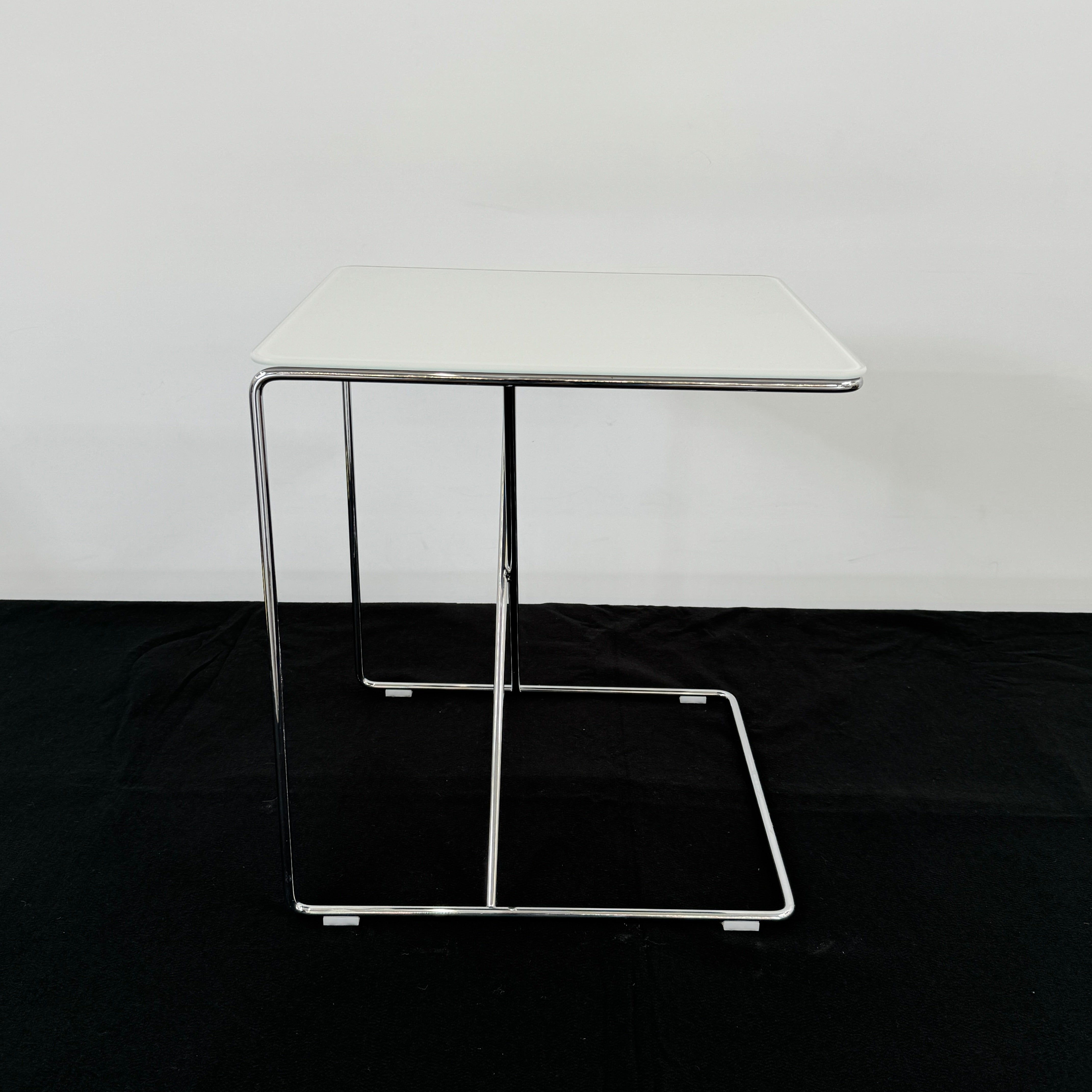 The B&B Italia JJ Cantilever Accent Table by B&B Italia features a white rectangular top and minimalist chrome frame, ideal for modern spaces. Shown against a plain white wall and black floor.