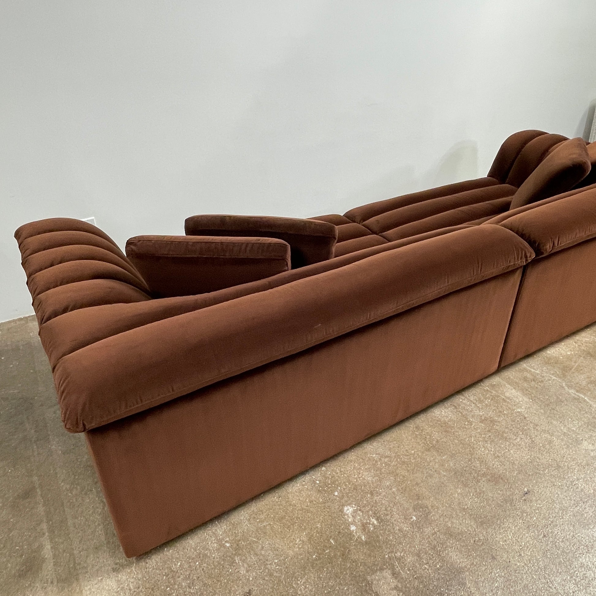 The Ellison Studios Float Sofa is a dark brown sectional from Ellison Studios with velvet upholstery, low profile, channel tufting, and two large cushions for extra comfort.