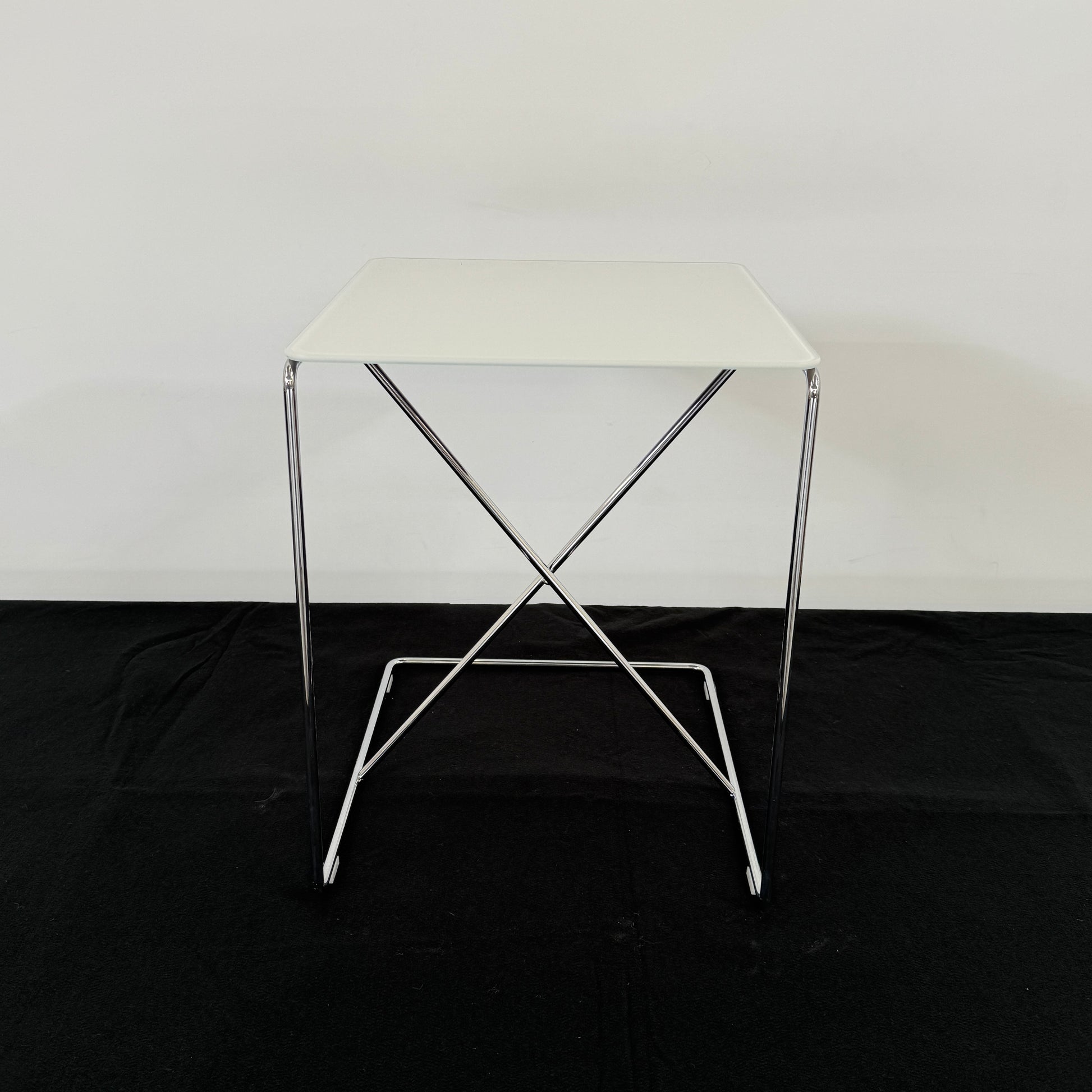 The B&B Italia JJ Cantilever Accent Table by B&B Italia features a white rectangular top and minimalist chrome frame, ideal for modern spaces. Shown against a plain white wall and black floor.