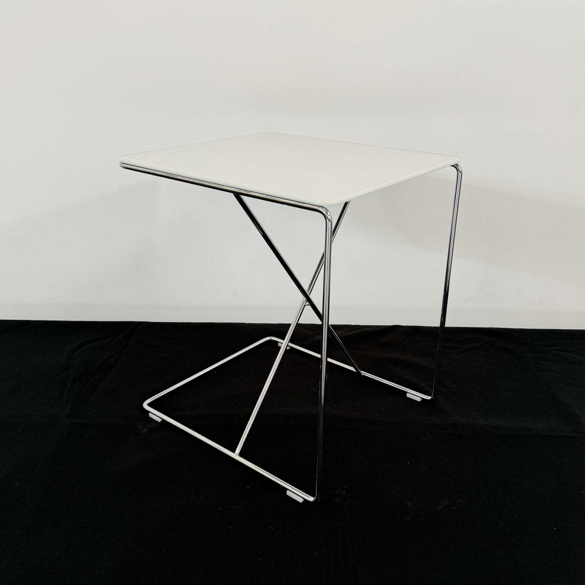 The B&B Italia JJ Cantilever Accent Table by B&B Italia features a white rectangular top and minimalist chrome frame, ideal for modern spaces. Shown against a plain white wall and black floor.