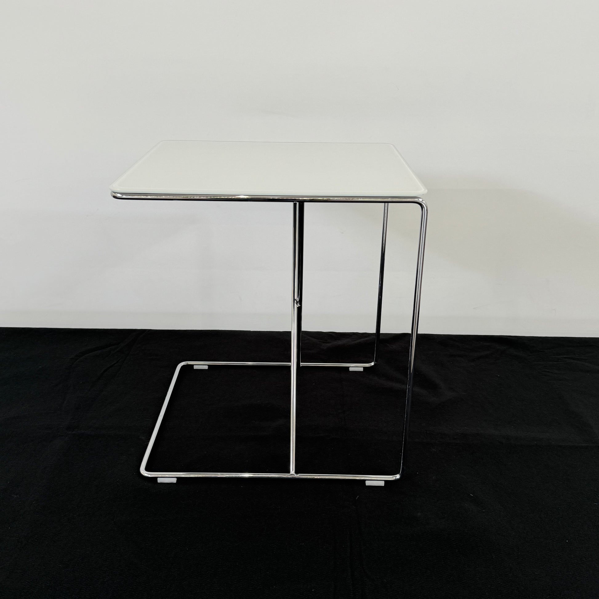 The B&B Italia JJ Cantilever Accent Table by B&B Italia features a white rectangular top and minimalist chrome frame, ideal for modern spaces. Shown against a plain white wall and black floor.
