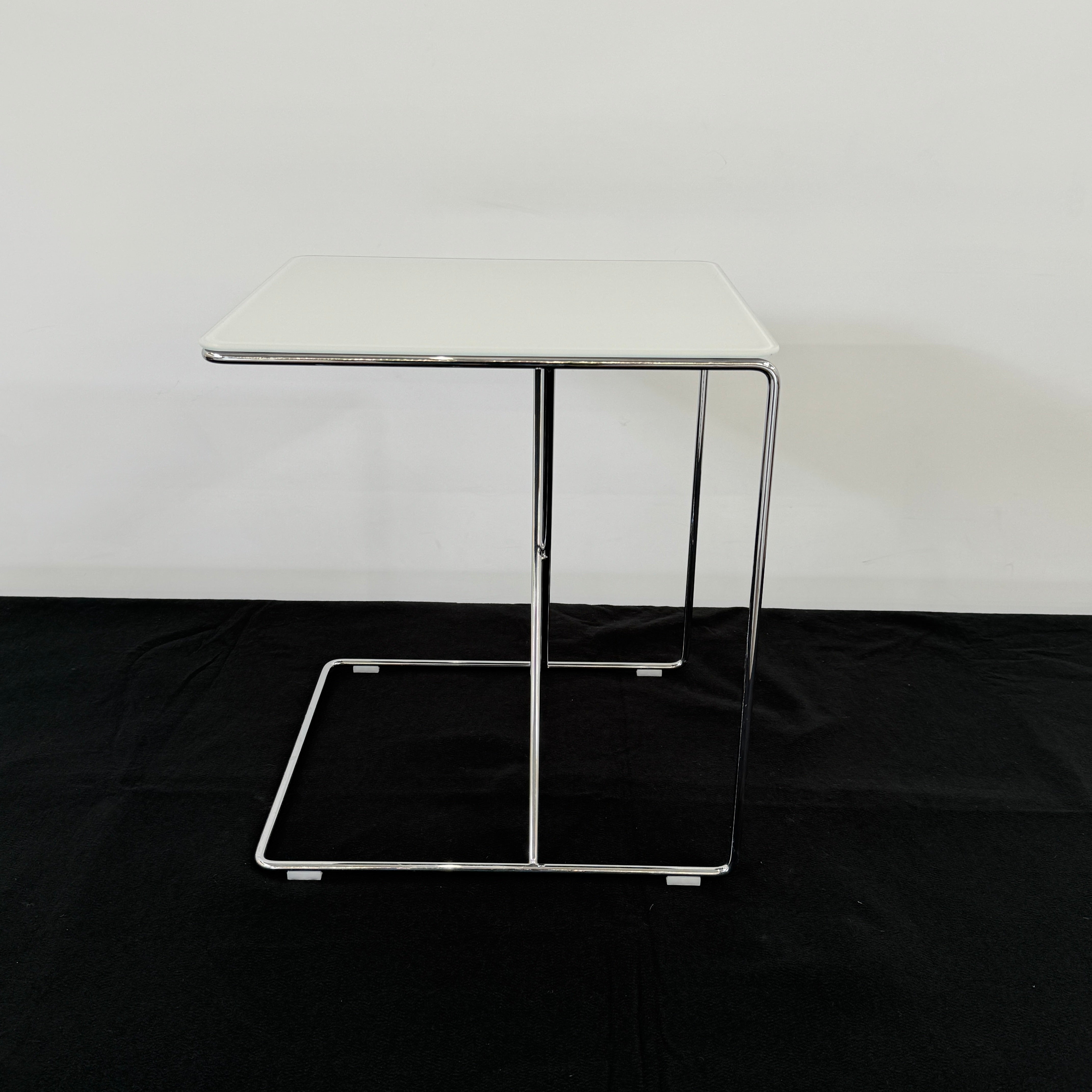 The B&B Italia JJ Cantilever Accent Table by B&B Italia features a white rectangular top and minimalist chrome frame, ideal for modern spaces. Shown against a plain white wall and black floor.