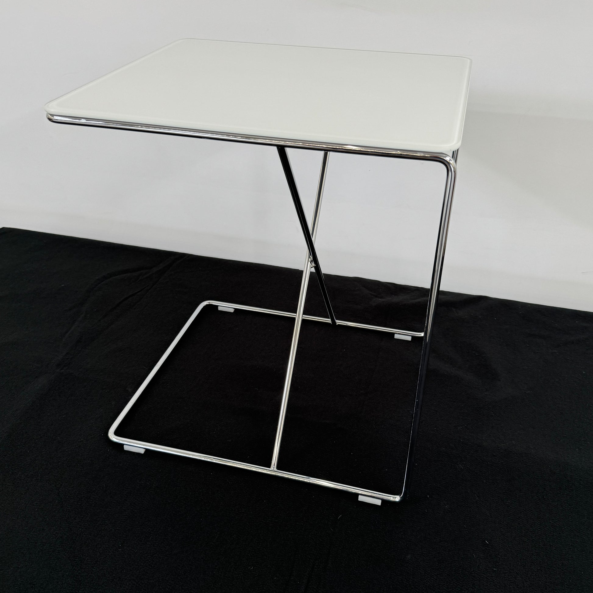 The B&B Italia JJ Cantilever Accent Table by B&B Italia features a white rectangular top and minimalist chrome frame, ideal for modern spaces. Shown against a plain white wall and black floor.