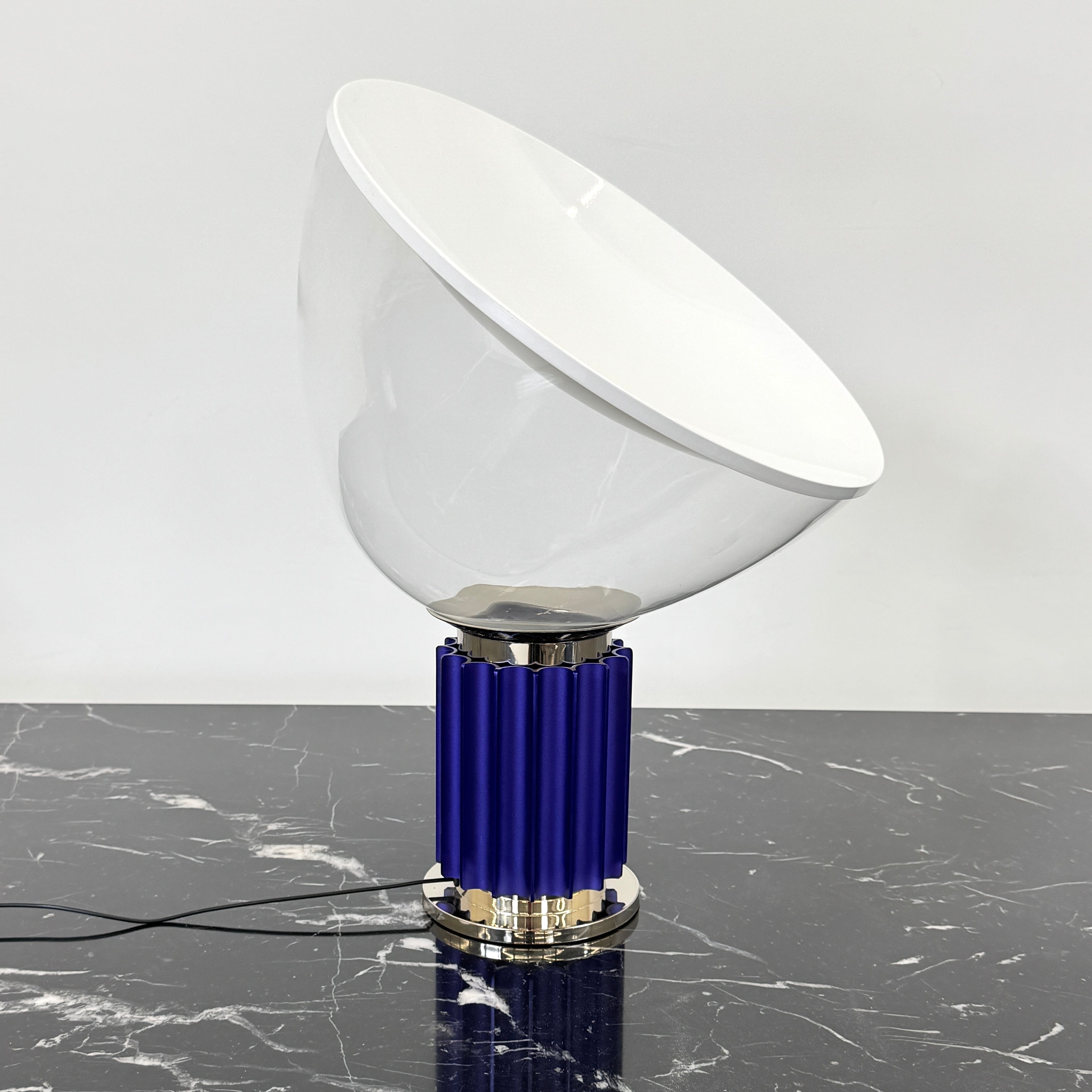 The Flos Taccia LED Table Lamp - Small (HOLD) by Flos, designed by Achille Castiglioni, features a large white bowl-shaped shade and a limited edition blue ribbed cylindrical base, shown against a plain grey background.