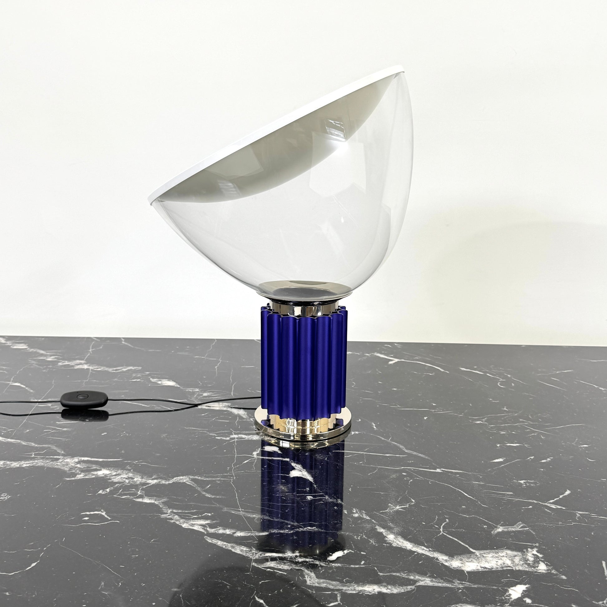 The Flos Taccia LED Table Lamp - Small (HOLD) by Flos, designed by Achille Castiglioni, features a large white bowl-shaped shade and a limited edition blue ribbed cylindrical base, shown against a plain grey background.