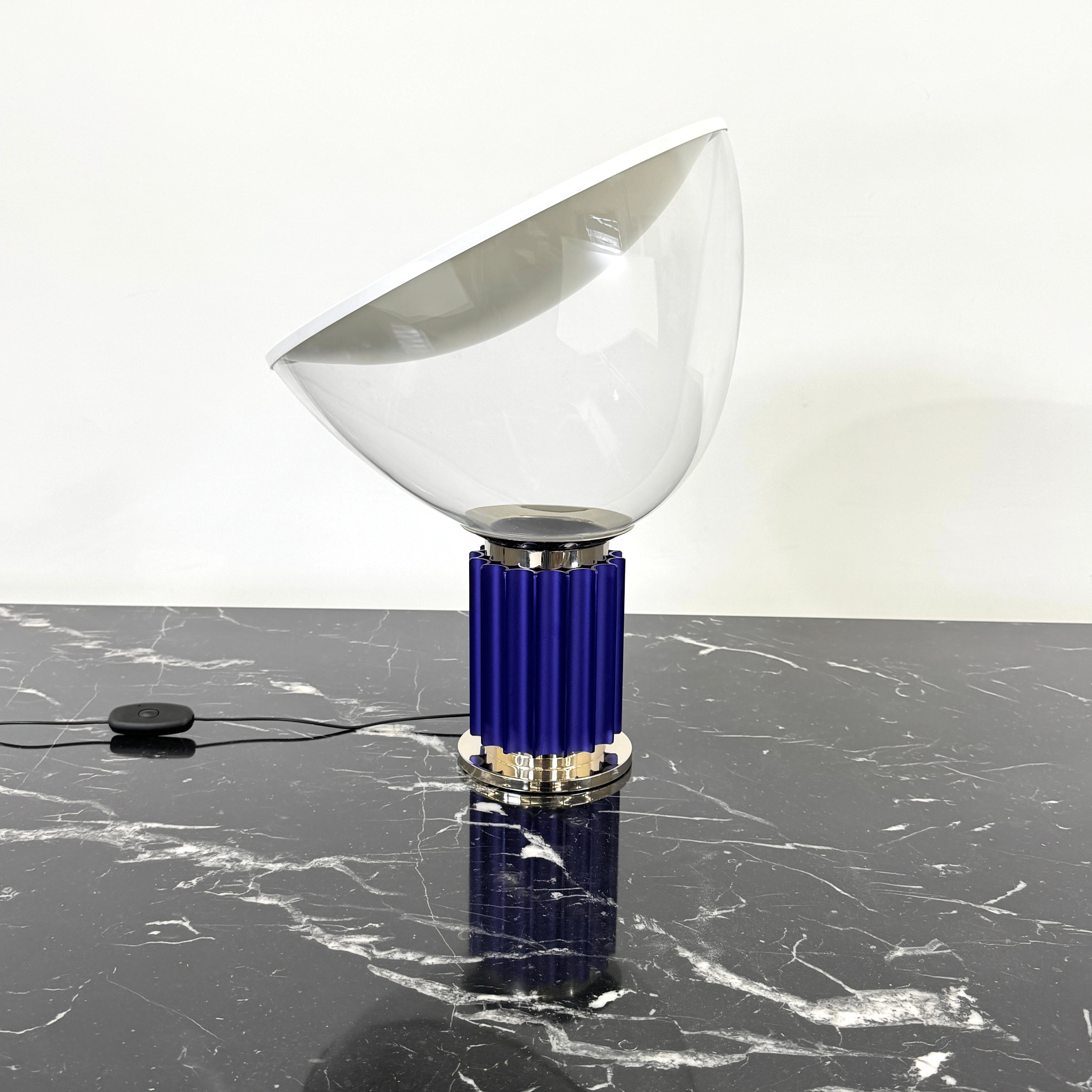 The Flos Taccia LED Table Lamp - Small (HOLD) by Flos, designed by Achille Castiglioni, features a large white bowl-shaped shade and a limited edition blue ribbed cylindrical base, shown against a plain grey background.