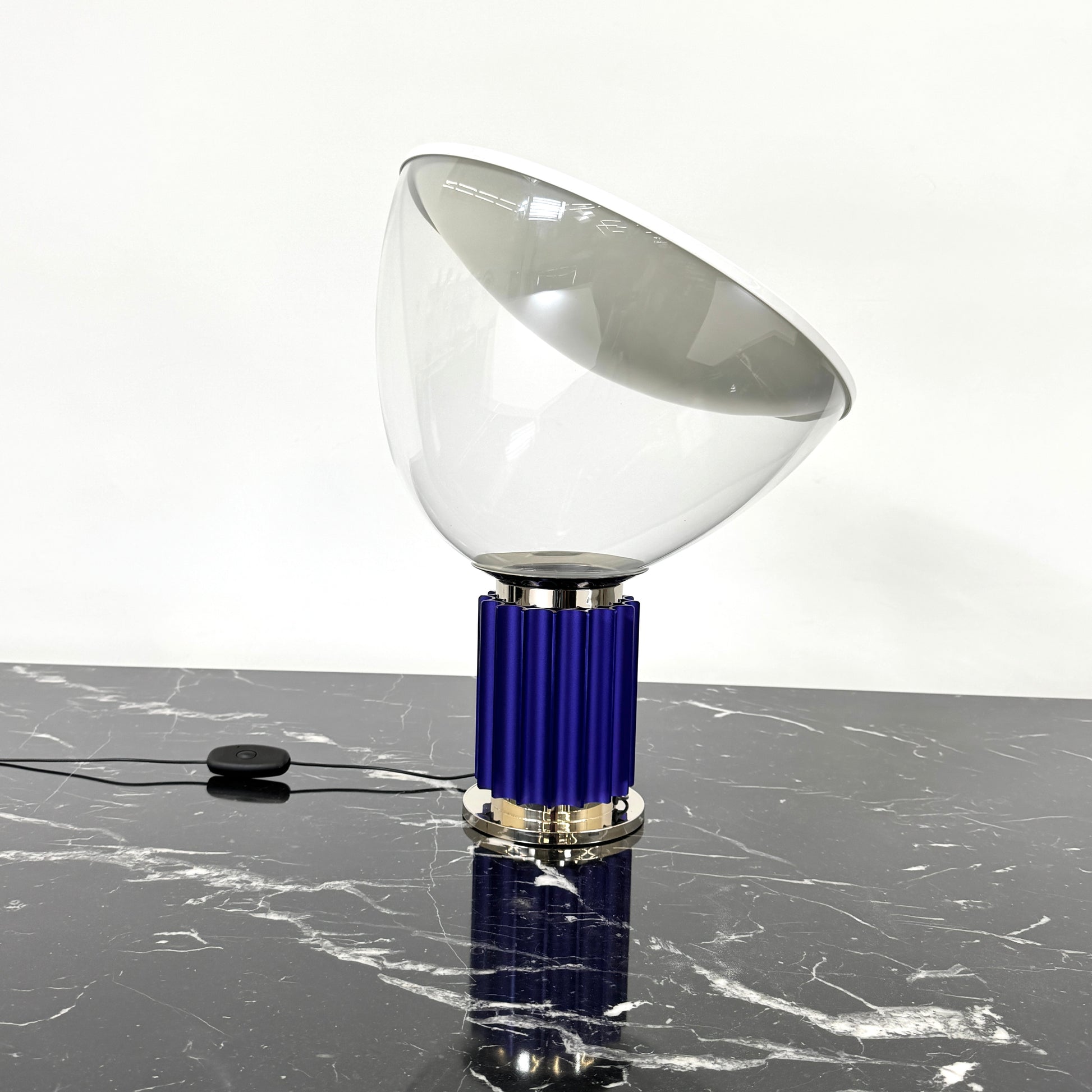 The Flos Taccia LED Table Lamp - Small (HOLD) by Flos, designed by Achille Castiglioni, features a large white bowl-shaped shade and a limited edition blue ribbed cylindrical base, shown against a plain grey background.