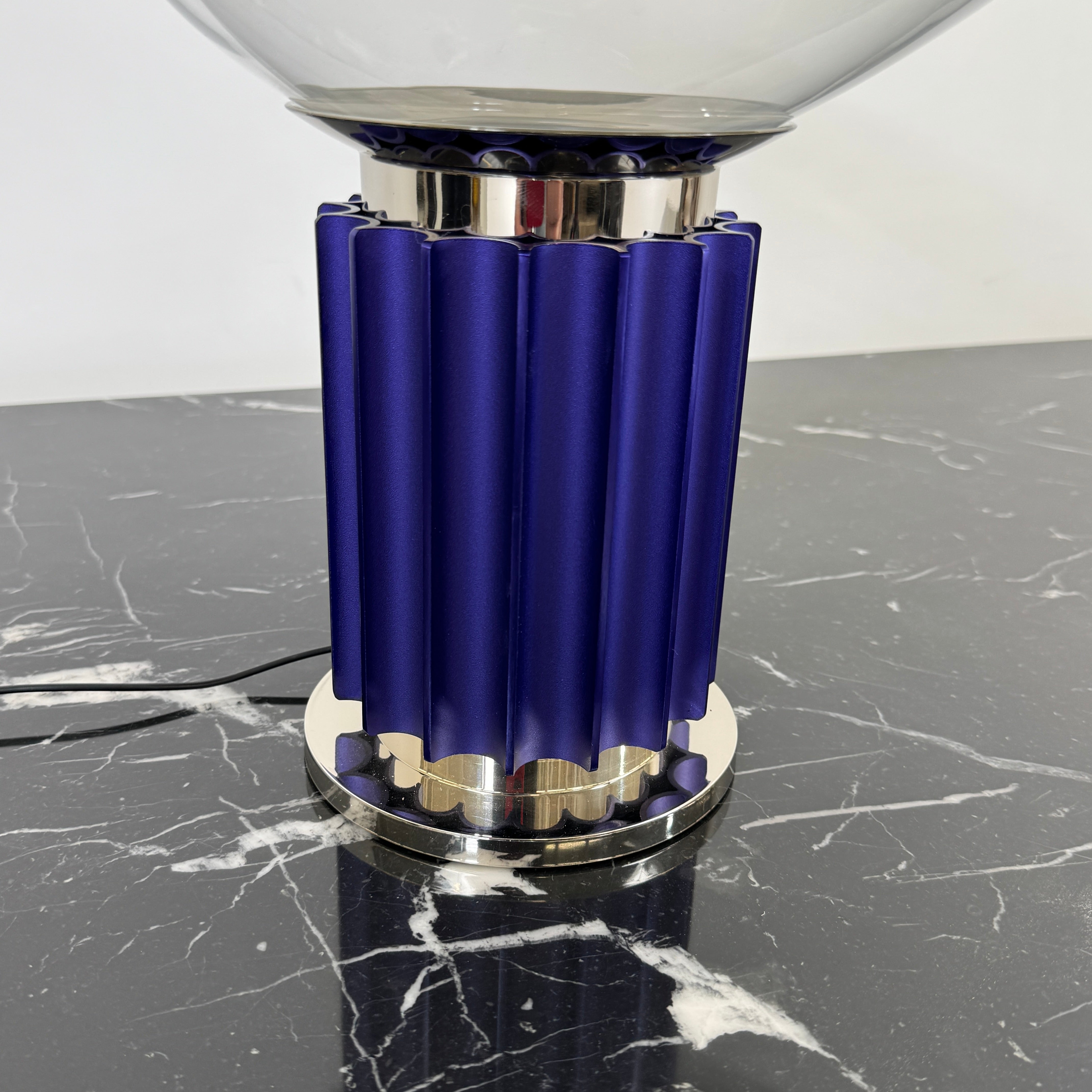 The Flos Taccia LED Table Lamp - Small (HOLD) by Flos, designed by Achille Castiglioni, features a large white bowl-shaped shade and a limited edition blue ribbed cylindrical base, shown against a plain grey background.