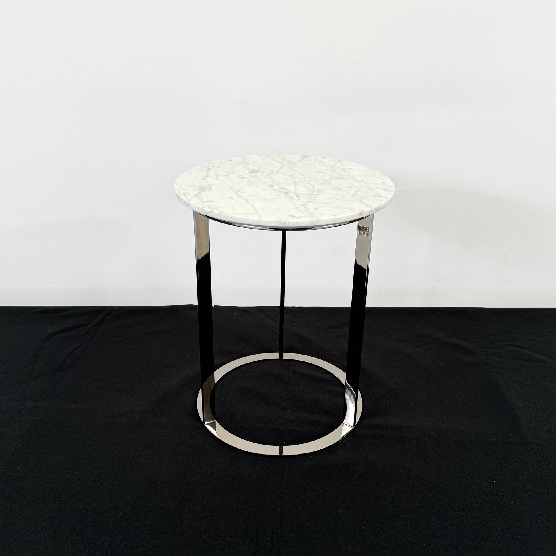 The B&B Italia Mera Accent Table features a round Carrara marble top and a three-legged metal frame with a circular base, showcasing B&B Italia's signature elegance.