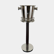 The Alessi Ettore Sottsass Wine Cooler and Stand features a stainless steel cooler with handles, mounted on a tall circular-base stand, and is set against a plain background.