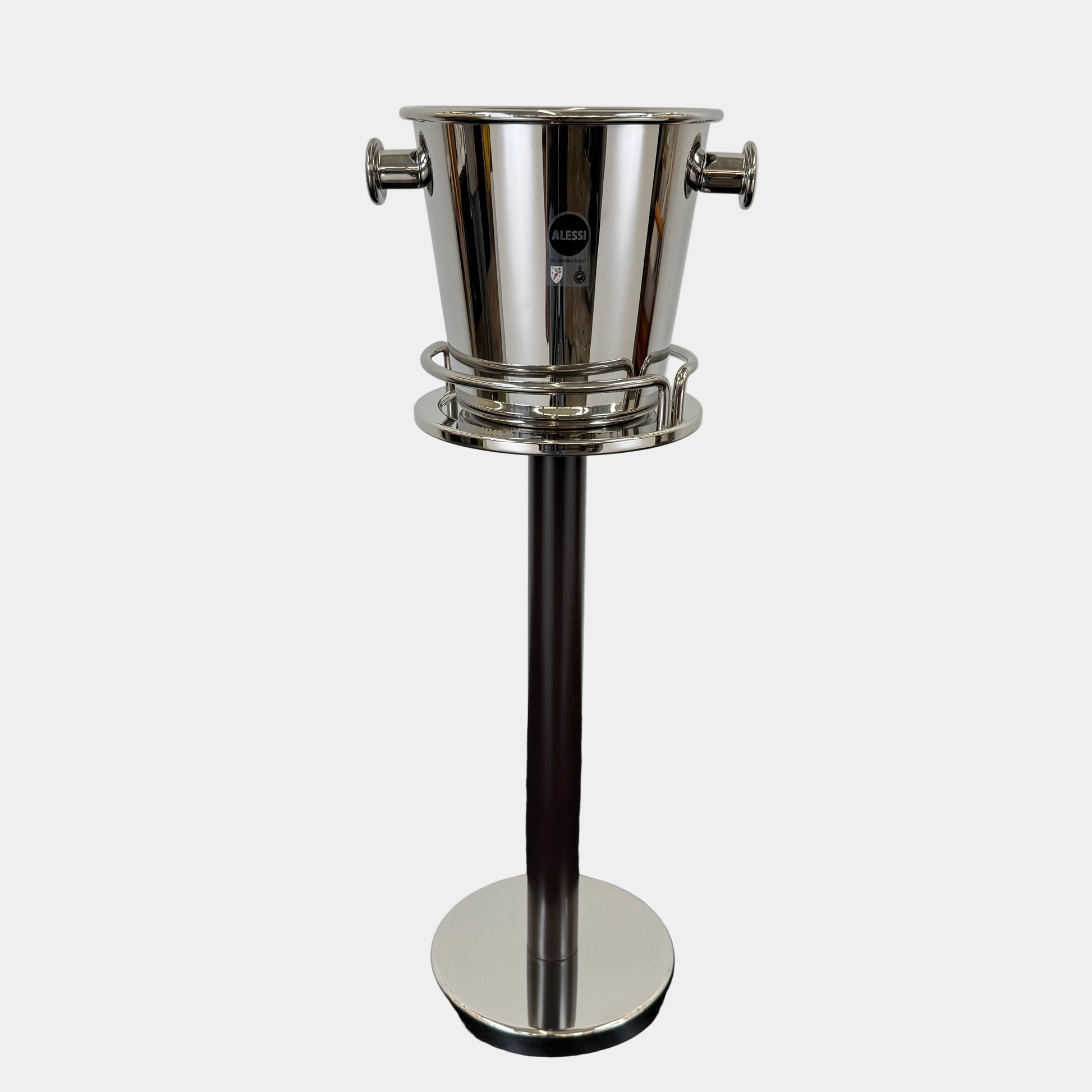 The Alessi Ettore Sottsass Wine Cooler and Stand features a stainless steel cooler with handles, mounted on a tall circular-base stand, and is set against a plain background.