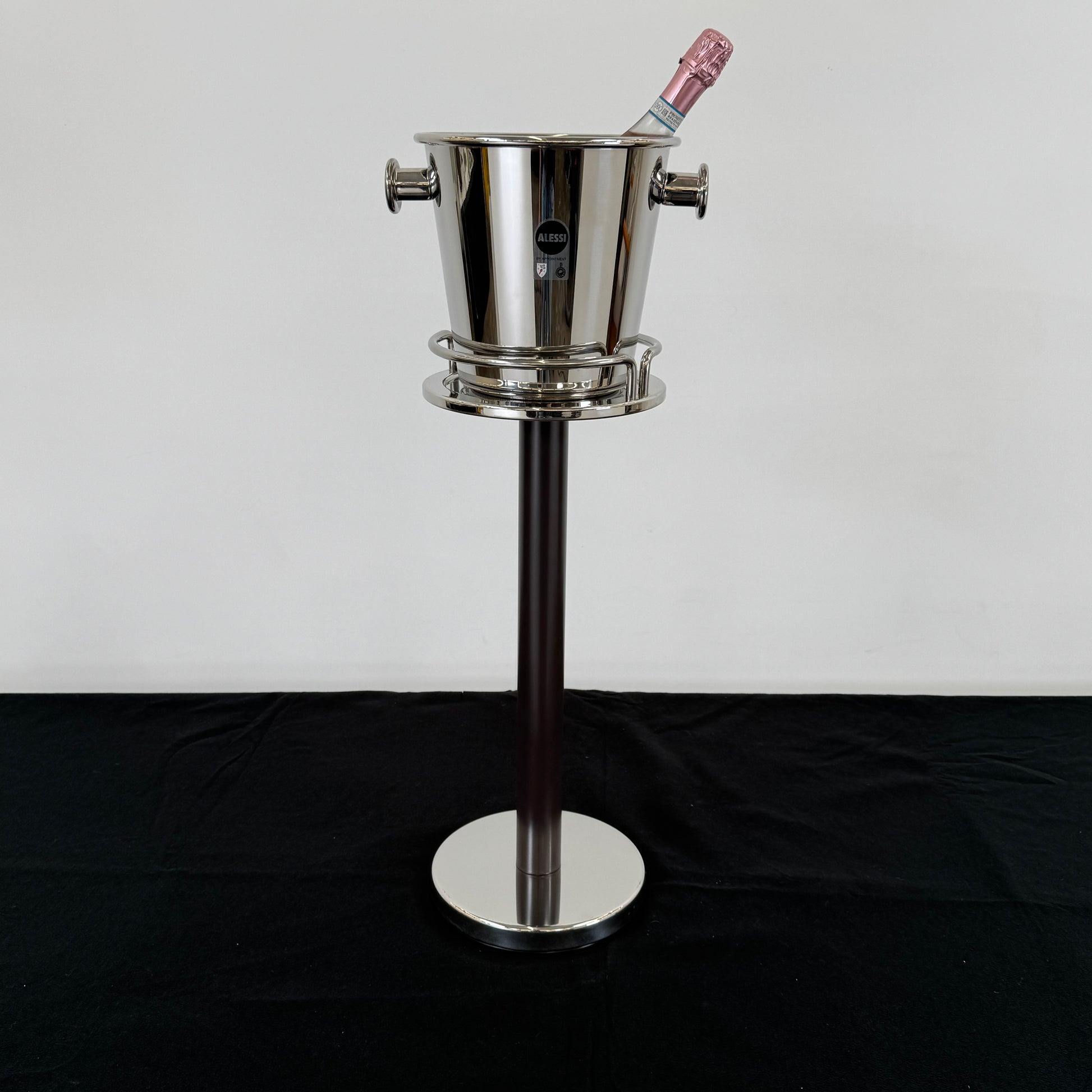The Alessi Ettore Sottsass Wine Cooler and Stand features a stainless steel cooler with handles, mounted on a tall circular-base stand, and is set against a plain background.