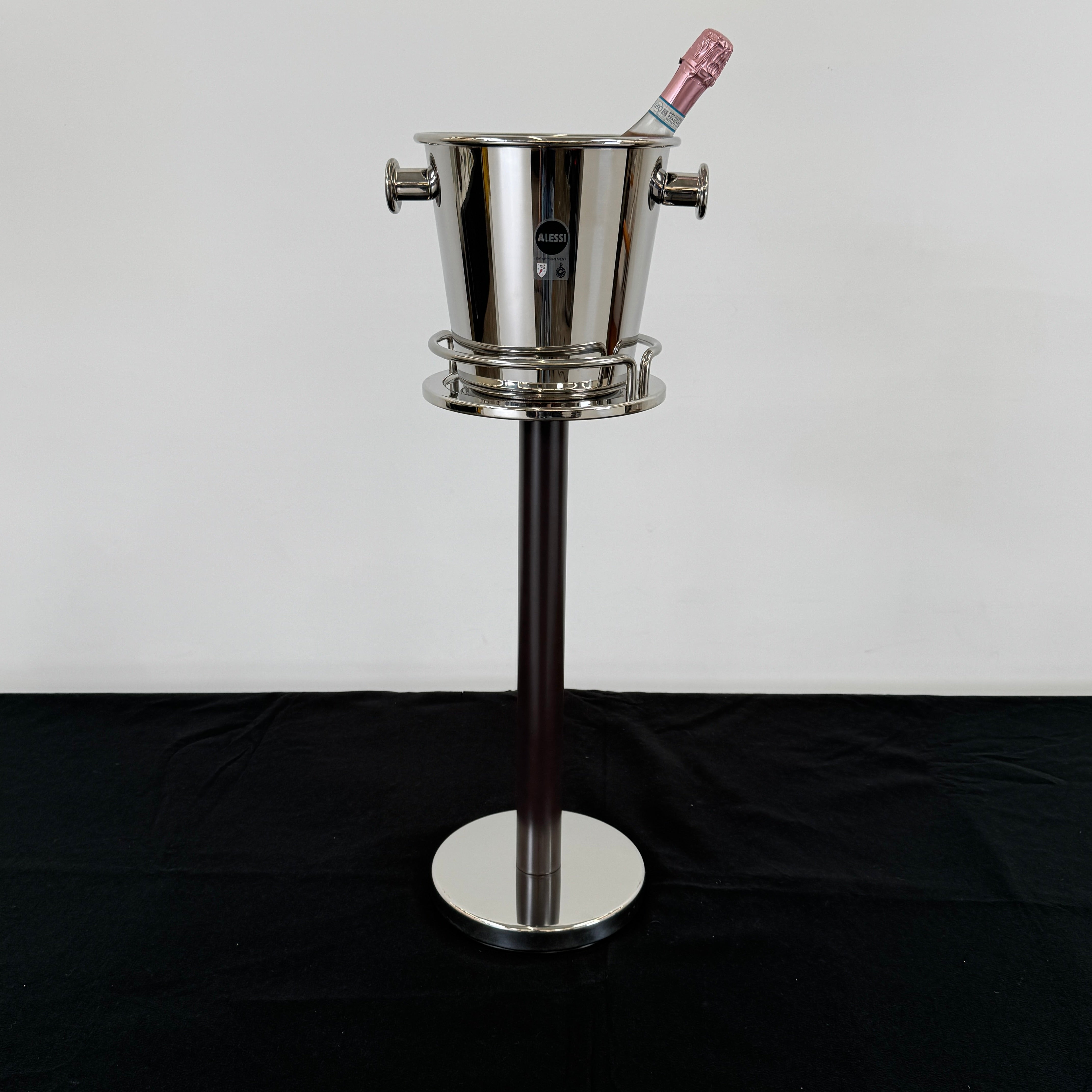 The Alessi Ettore Sottsass Wine Cooler and Stand features a stainless steel cooler with handles, mounted on a tall circular-base stand, and is set against a plain background.