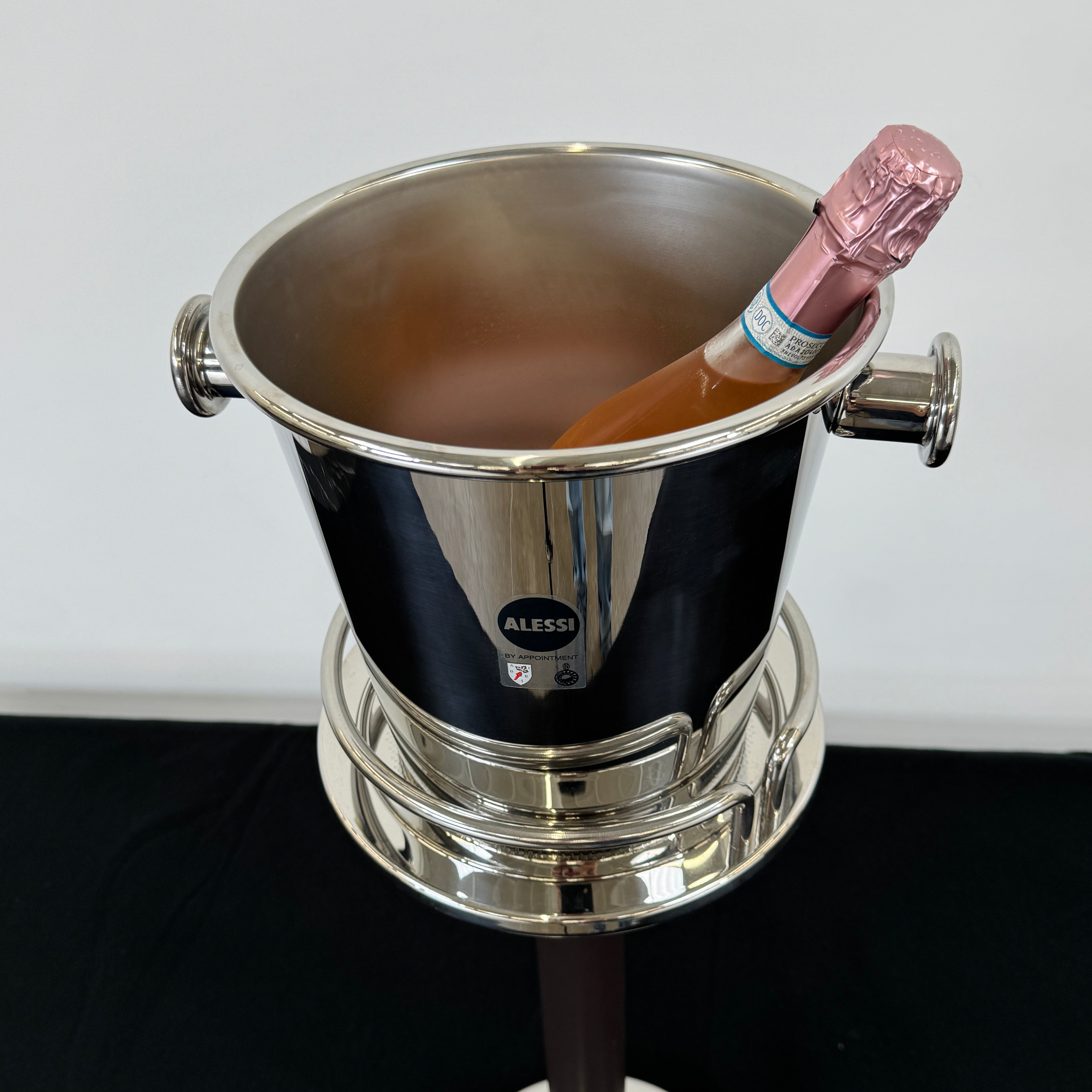 The Alessi Ettore Sottsass Wine Cooler and Stand features a stainless steel cooler with handles, mounted on a tall circular-base stand, and is set against a plain background.