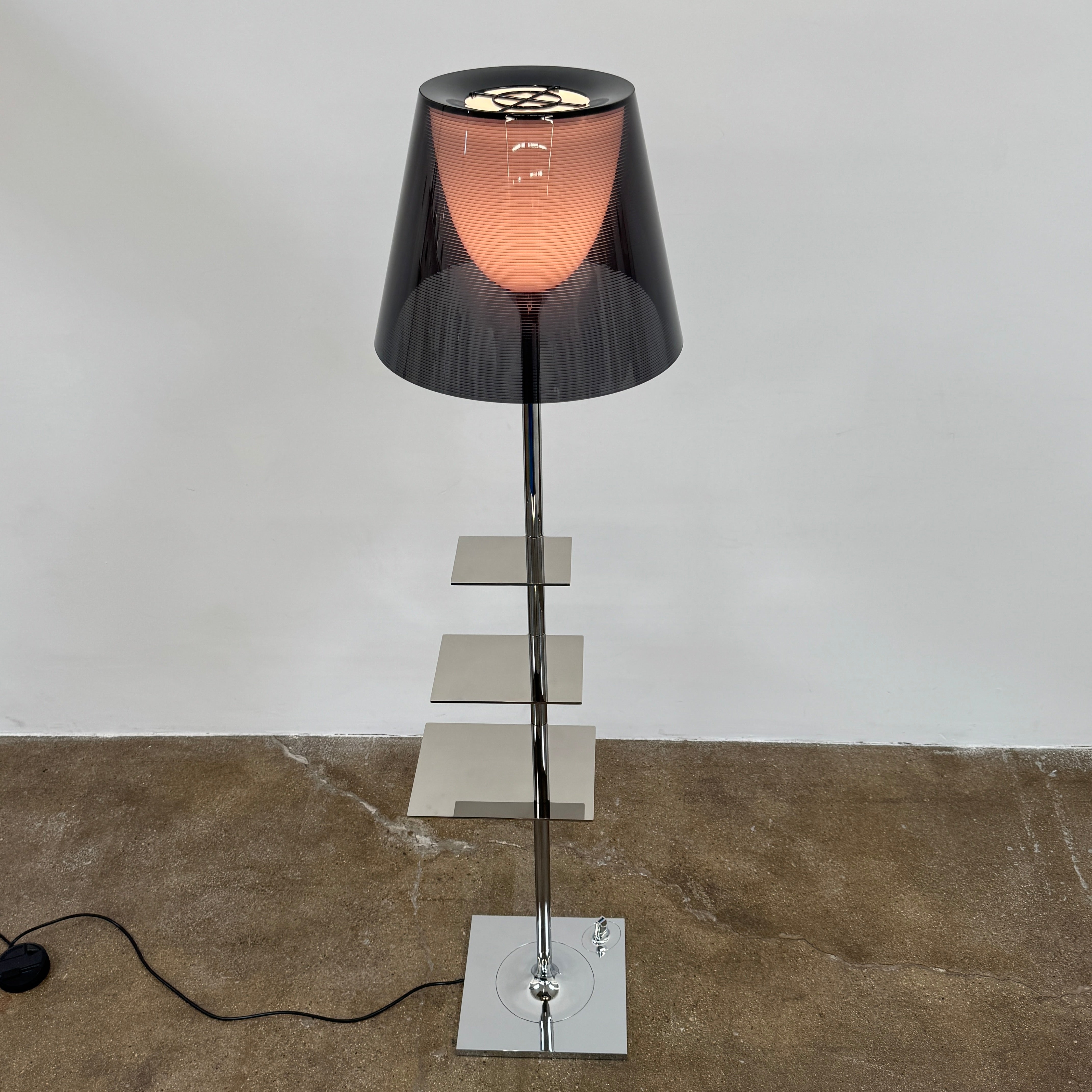 The Flos Bibliotheque Floor Lamp by Flos features a black translucent shade, metal stand, three rotating shelves, USB charging port, and square base, standing on a brown floor against a white wall.