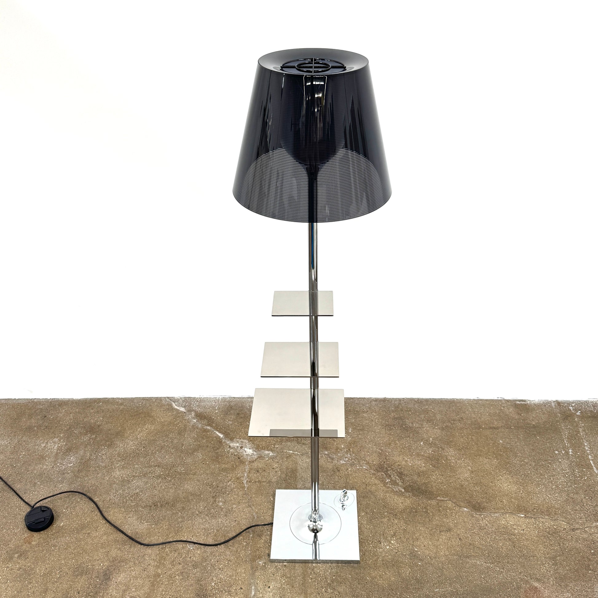The Flos Bibliotheque Floor Lamp by Flos features a black translucent shade, metal stand, three rotating shelves, USB charging port, and square base, standing on a brown floor against a white wall.
