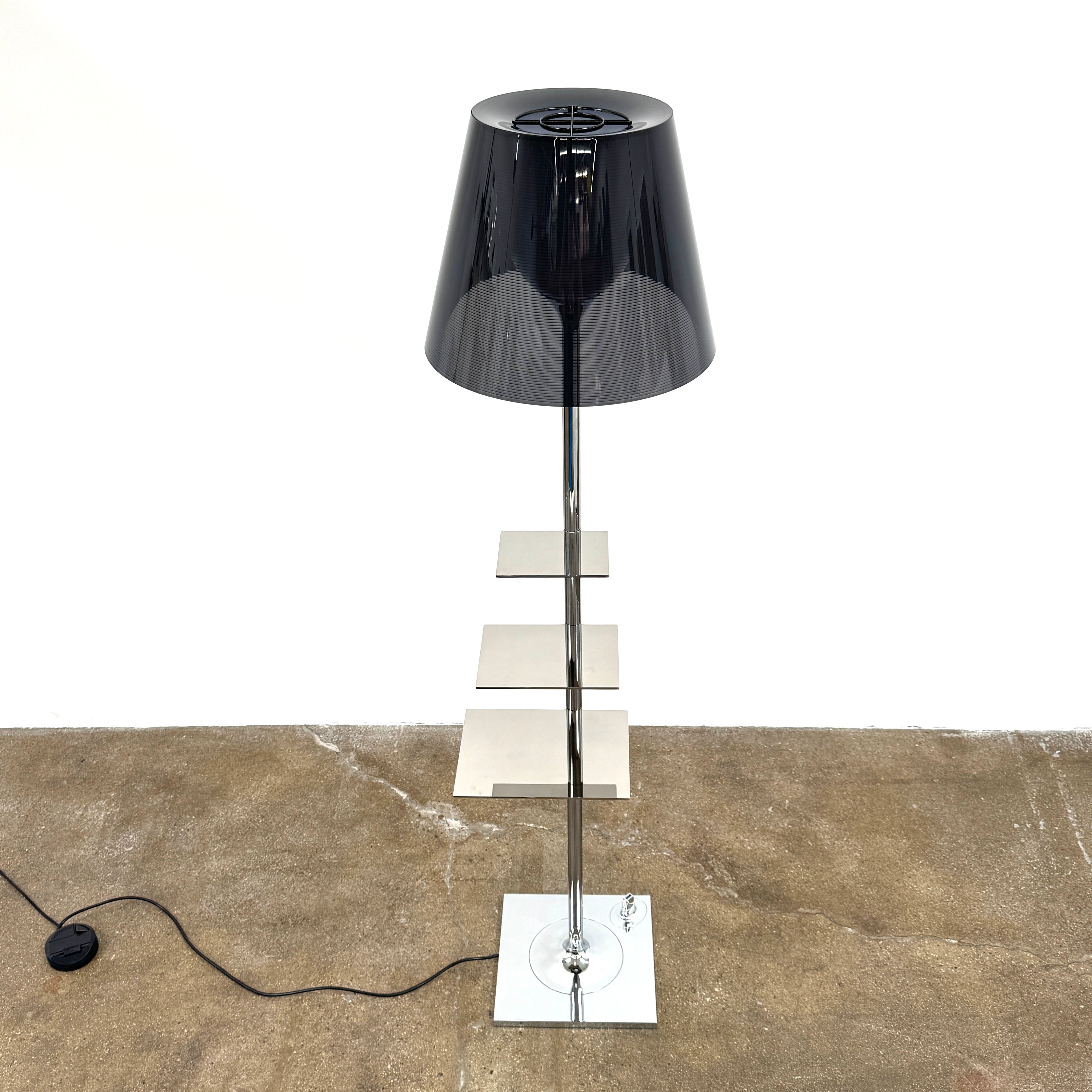 The Flos Bibliotheque Floor Lamp by Flos features a black translucent shade, metal stand, three rotating shelves, USB charging port, and square base, standing on a brown floor against a white wall.