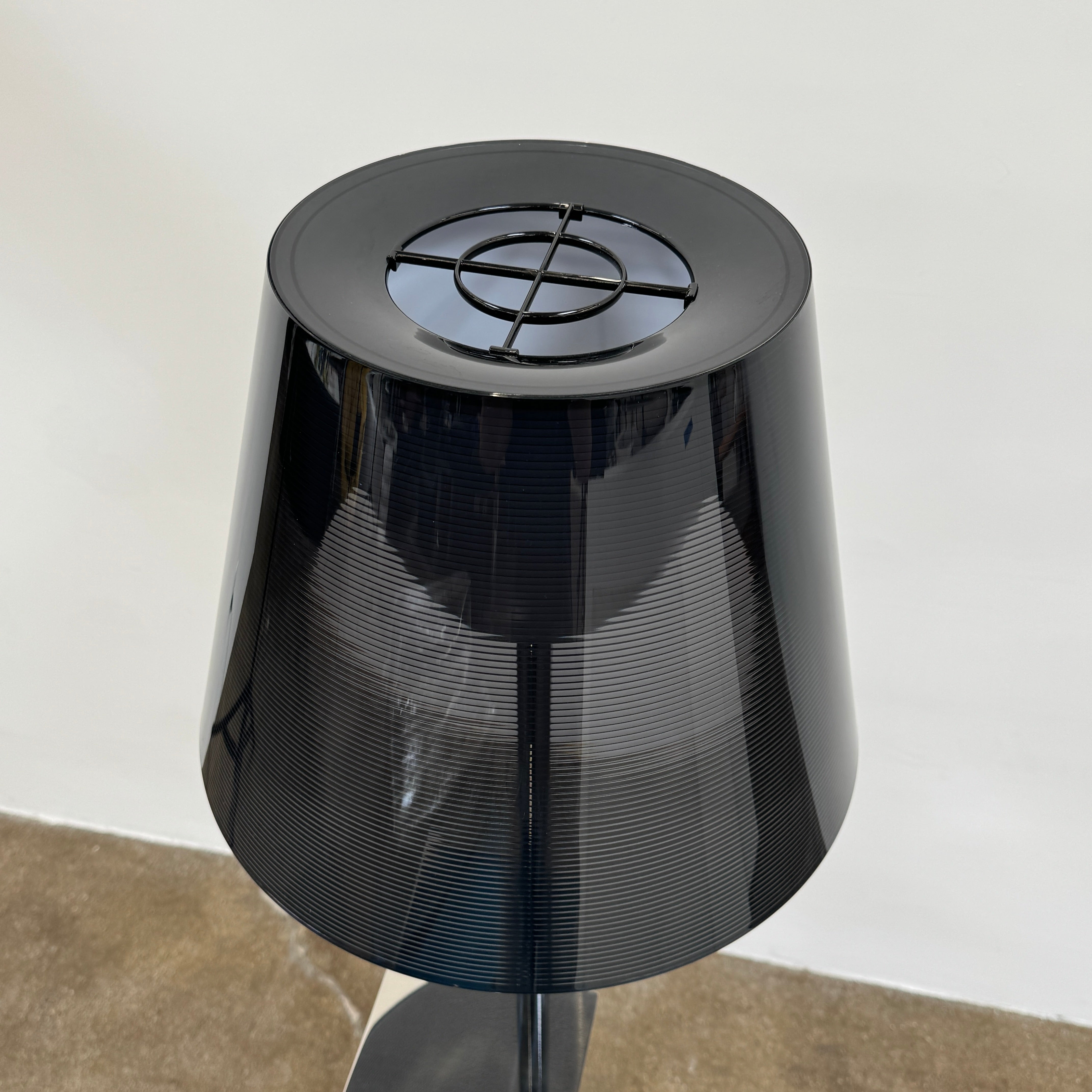 The Flos Bibliotheque Floor Lamp by Flos features a black translucent shade, metal stand, three rotating shelves, USB charging port, and square base, standing on a brown floor against a white wall.