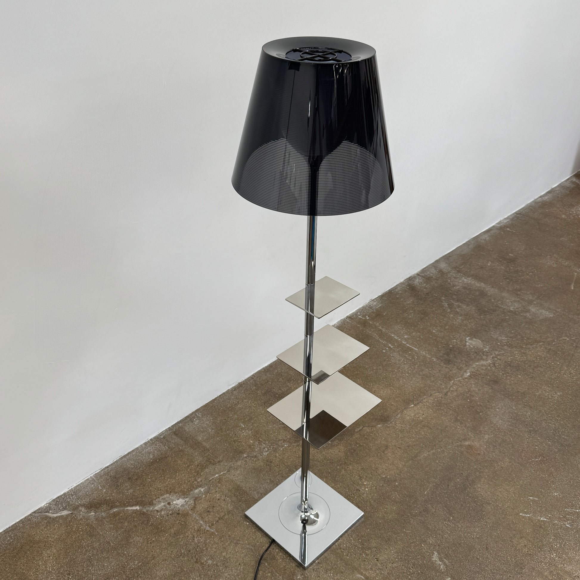 The Flos Bibliotheque Floor Lamp by Flos features a black translucent shade, metal stand, three rotating shelves, USB charging port, and square base, standing on a brown floor against a white wall.