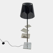 The Flos Bibliotheque Floor Lamp by Flos features a black translucent shade, metal stand, three rotating shelves, USB charging port, and square base, standing on a brown floor against a white wall.