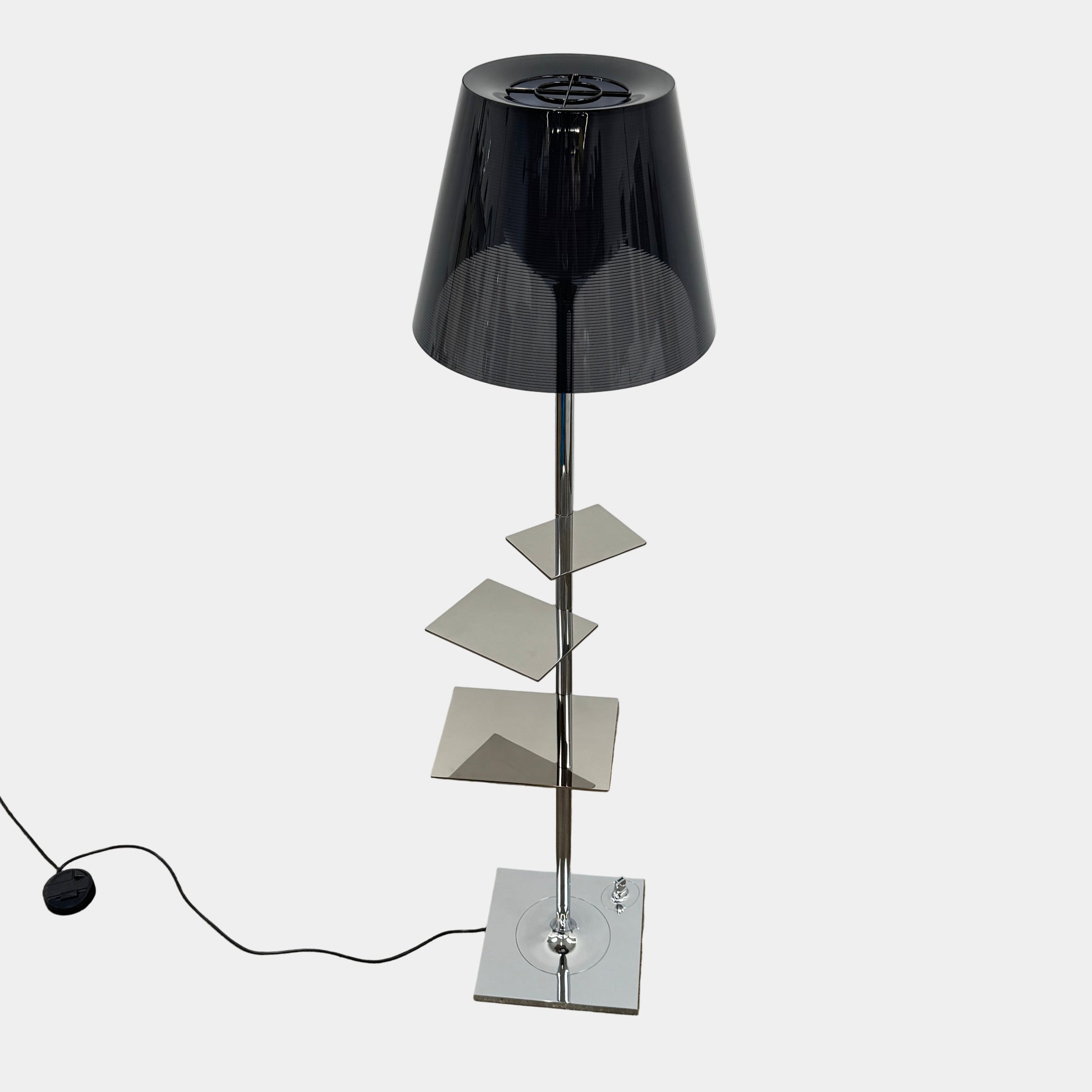 The Flos Bibliotheque Floor Lamp by Flos features a black translucent shade, metal stand, three rotating shelves, USB charging port, and square base, standing on a brown floor against a white wall.