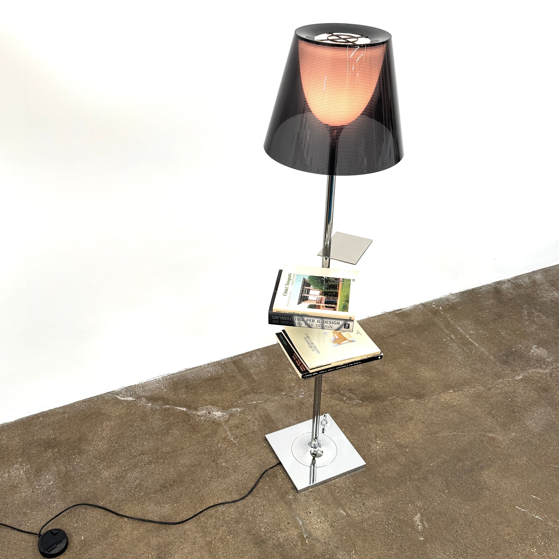 The Flos Bibliotheque Floor Lamp by Flos features a black translucent shade, metal stand, three rotating shelves, USB charging port, and square base, standing on a brown floor against a white wall.