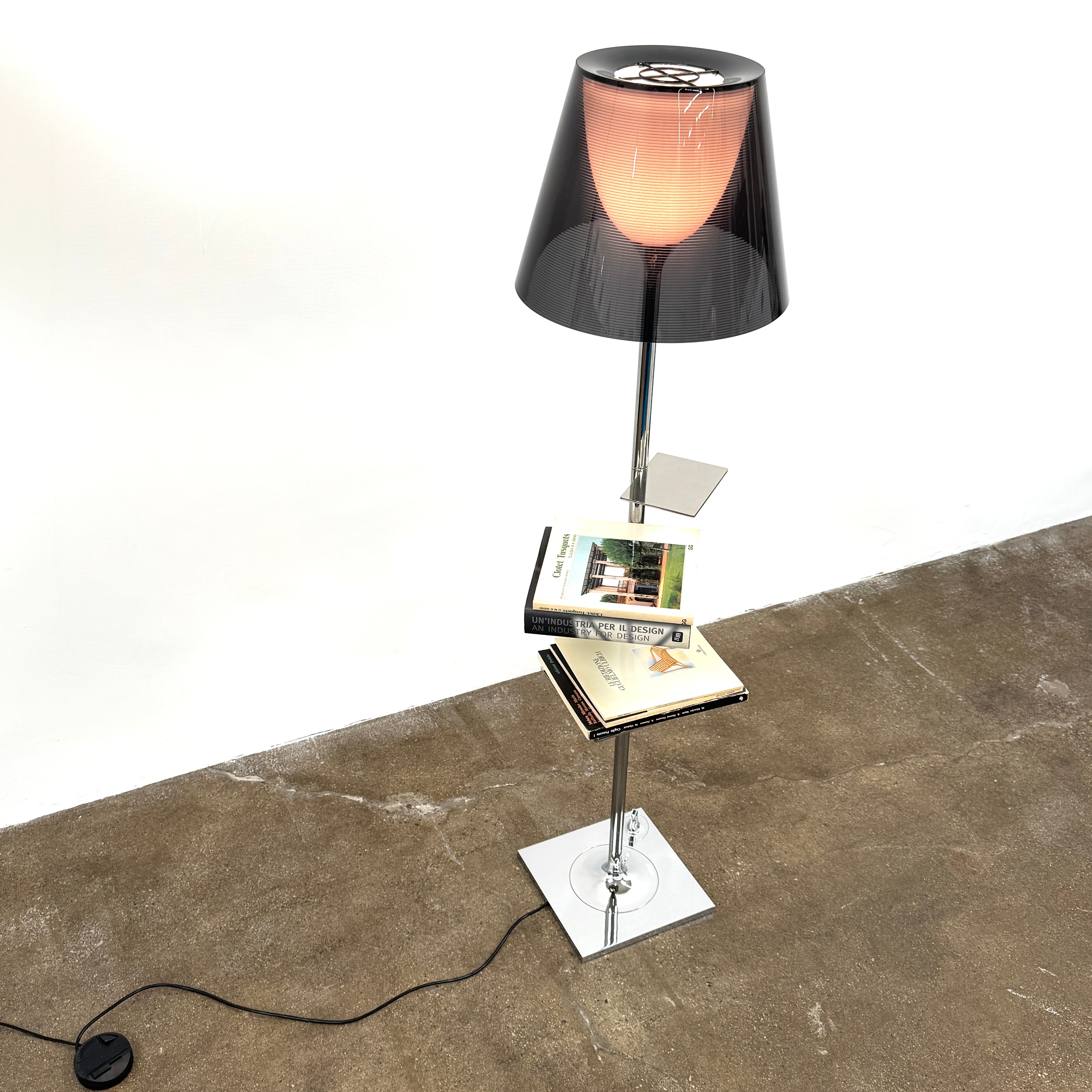 The Flos Bibliotheque Floor Lamp by Flos features a black translucent shade, metal stand, three rotating shelves, USB charging port, and square base, standing on a brown floor against a white wall.