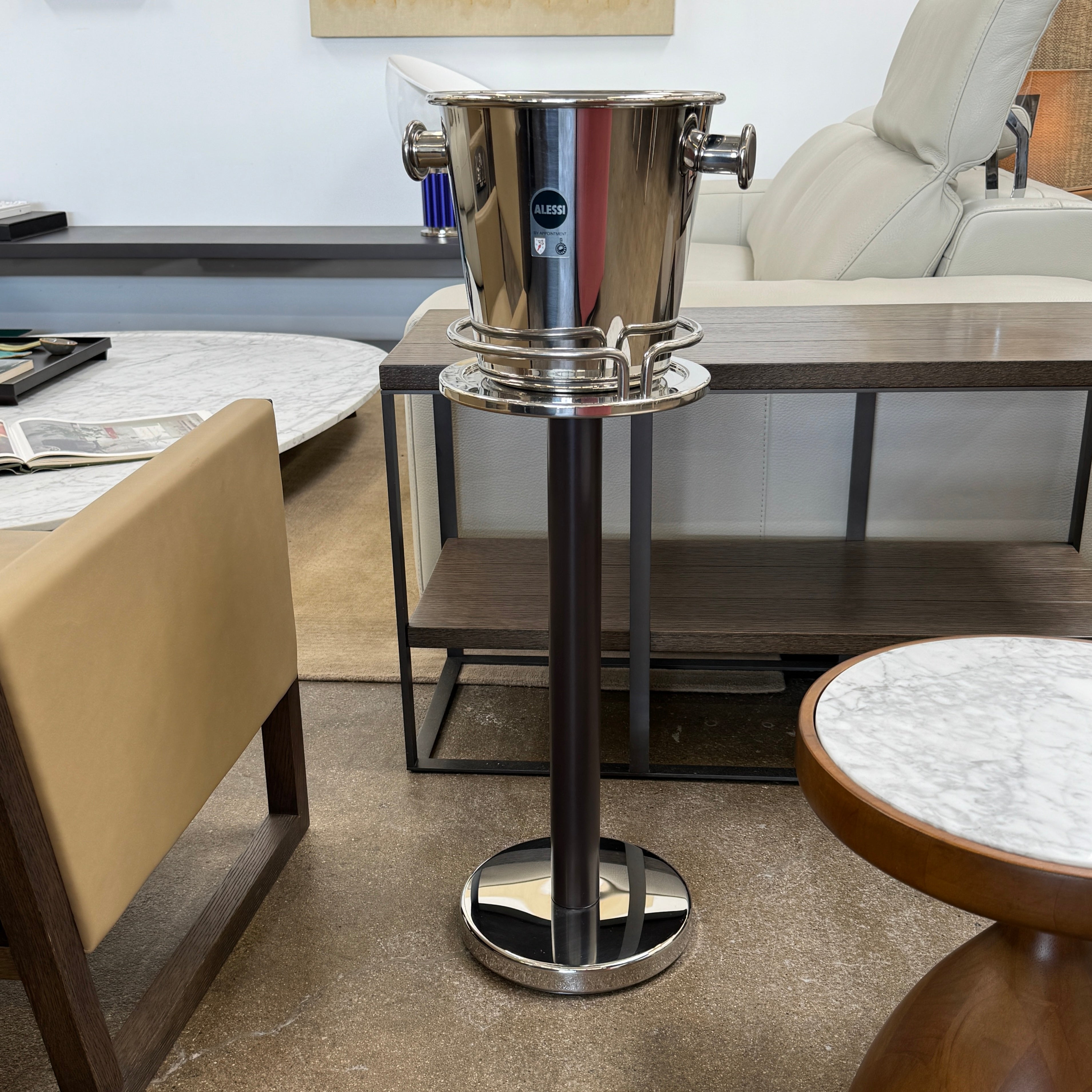 The Alessi Ettore Sottsass Wine Cooler and Stand features a stainless steel cooler with handles, mounted on a tall circular-base stand, and is set against a plain background.