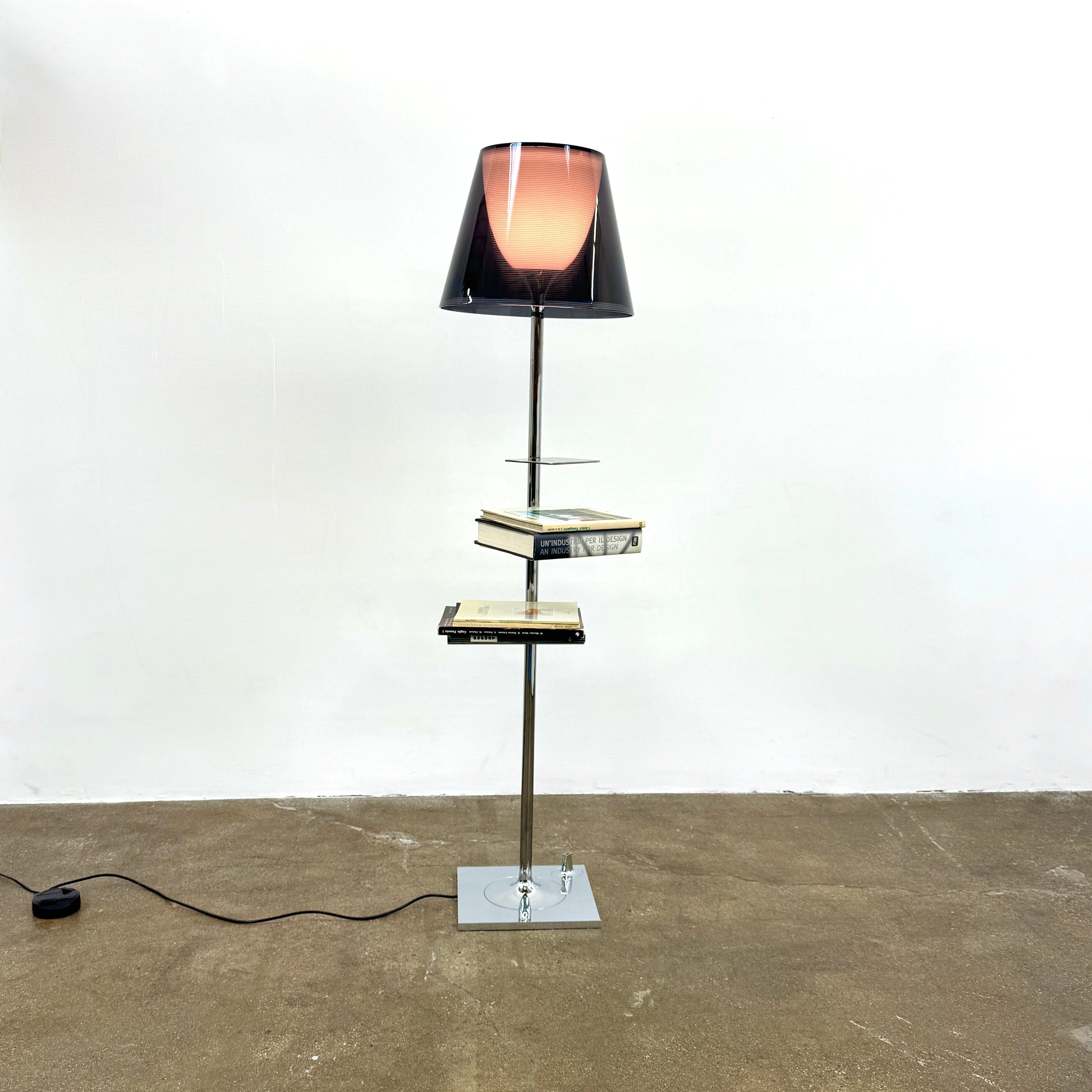 The Flos Bibliotheque Floor Lamp by Flos features a black translucent shade, metal stand, three rotating shelves, USB charging port, and square base, standing on a brown floor against a white wall.