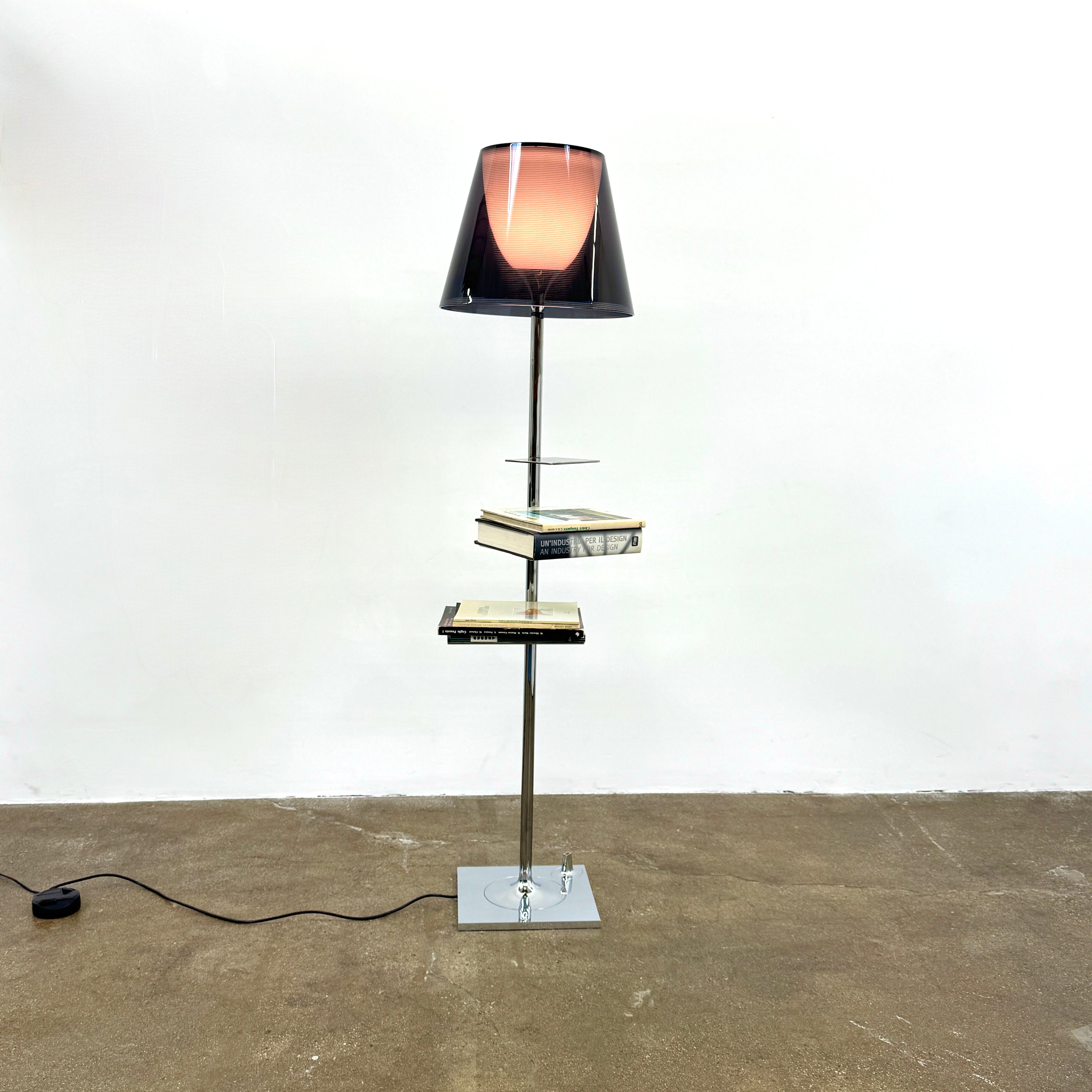 The Flos Bibliotheque Floor Lamp by Flos features a black translucent shade, metal stand, three rotating shelves, USB charging port, and square base, standing on a brown floor against a white wall.