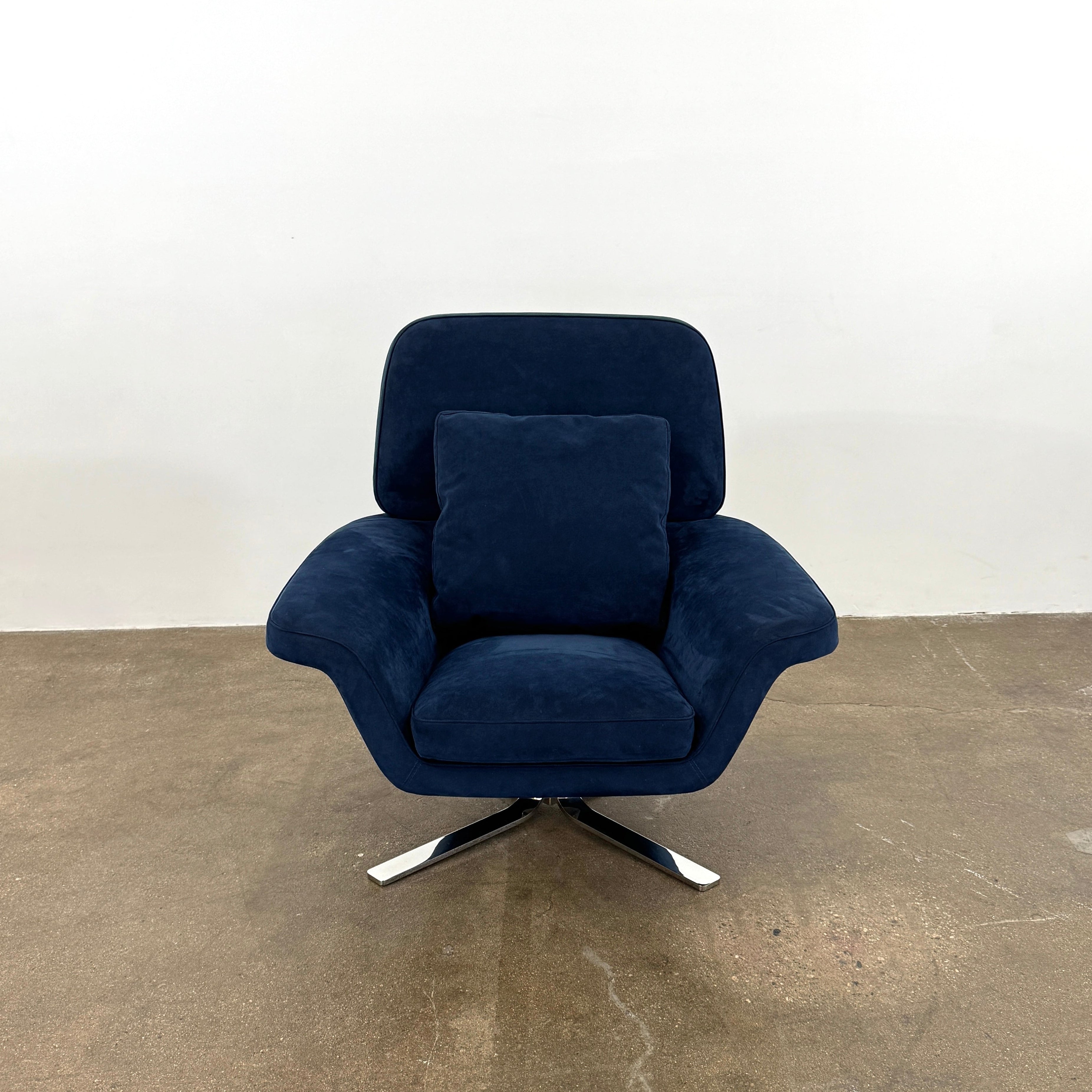 The Minotti Blake Swivel Armchair in modern navy blue, featuring a matching cushion and metal swivel base, is placed on a bare concrete floor against a plain white wall.