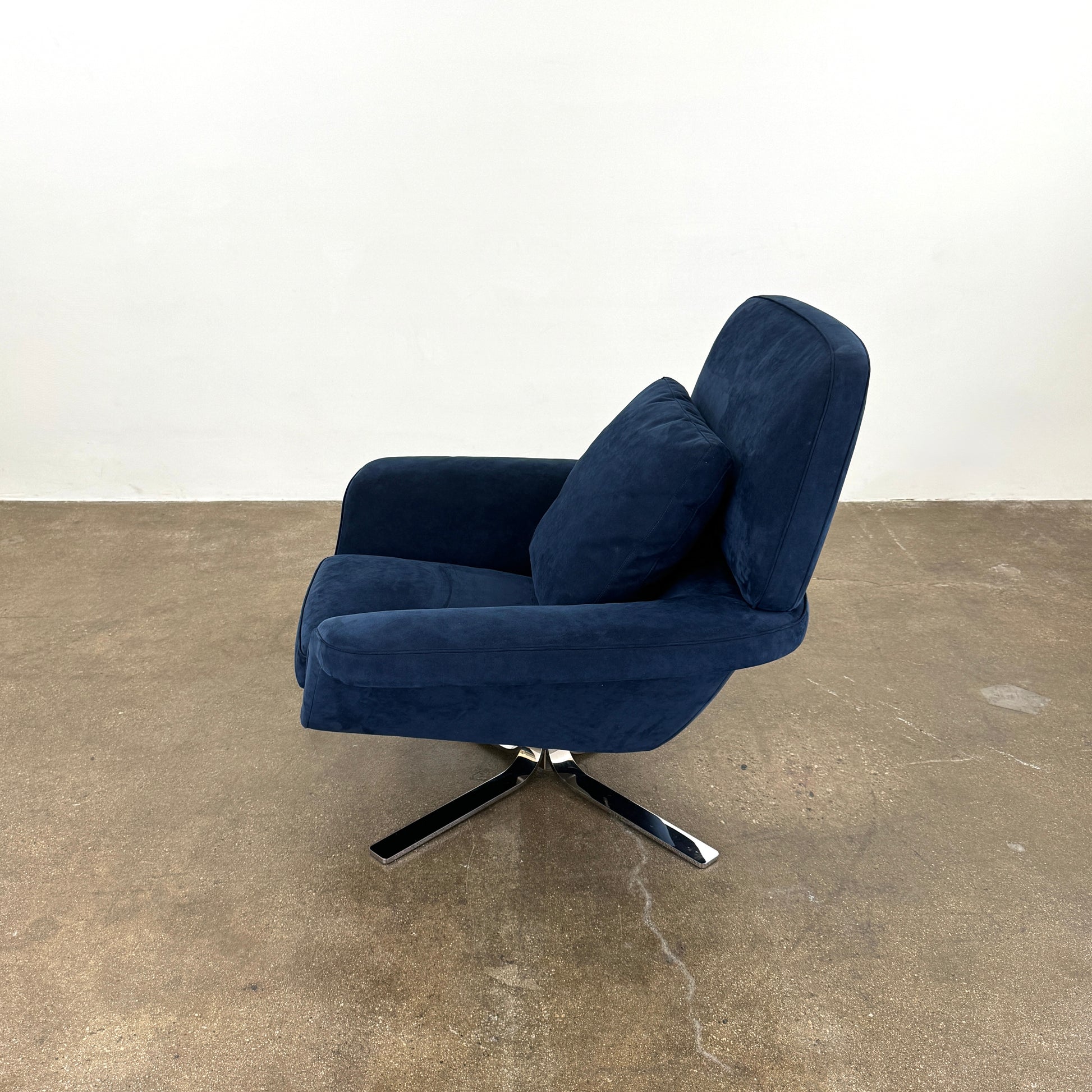 The Minotti Blake Swivel Armchair in modern navy blue, featuring a matching cushion and metal swivel base, is placed on a bare concrete floor against a plain white wall.