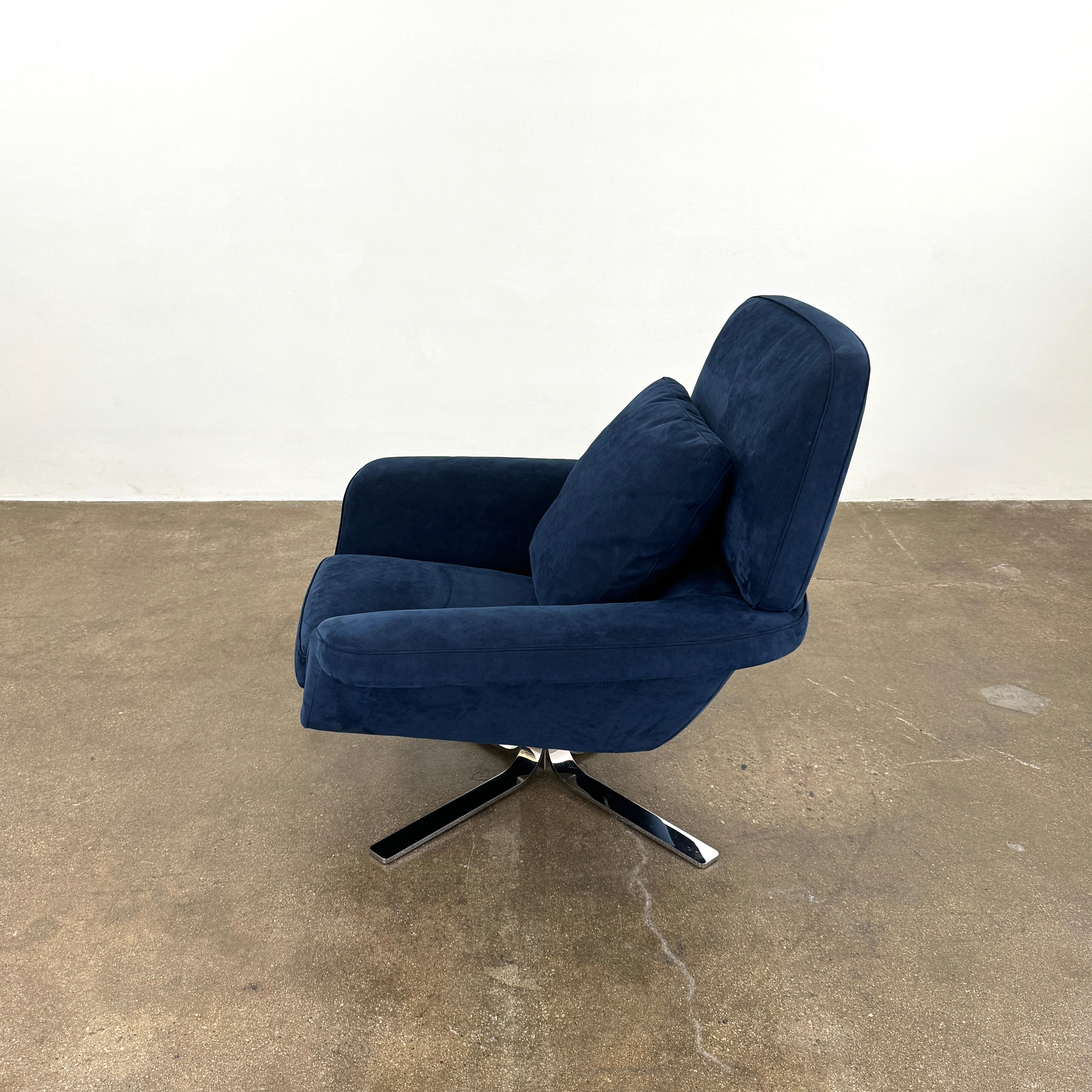 The Minotti Blake Swivel Armchair in modern navy blue, featuring a matching cushion and metal swivel base, is placed on a bare concrete floor against a plain white wall.