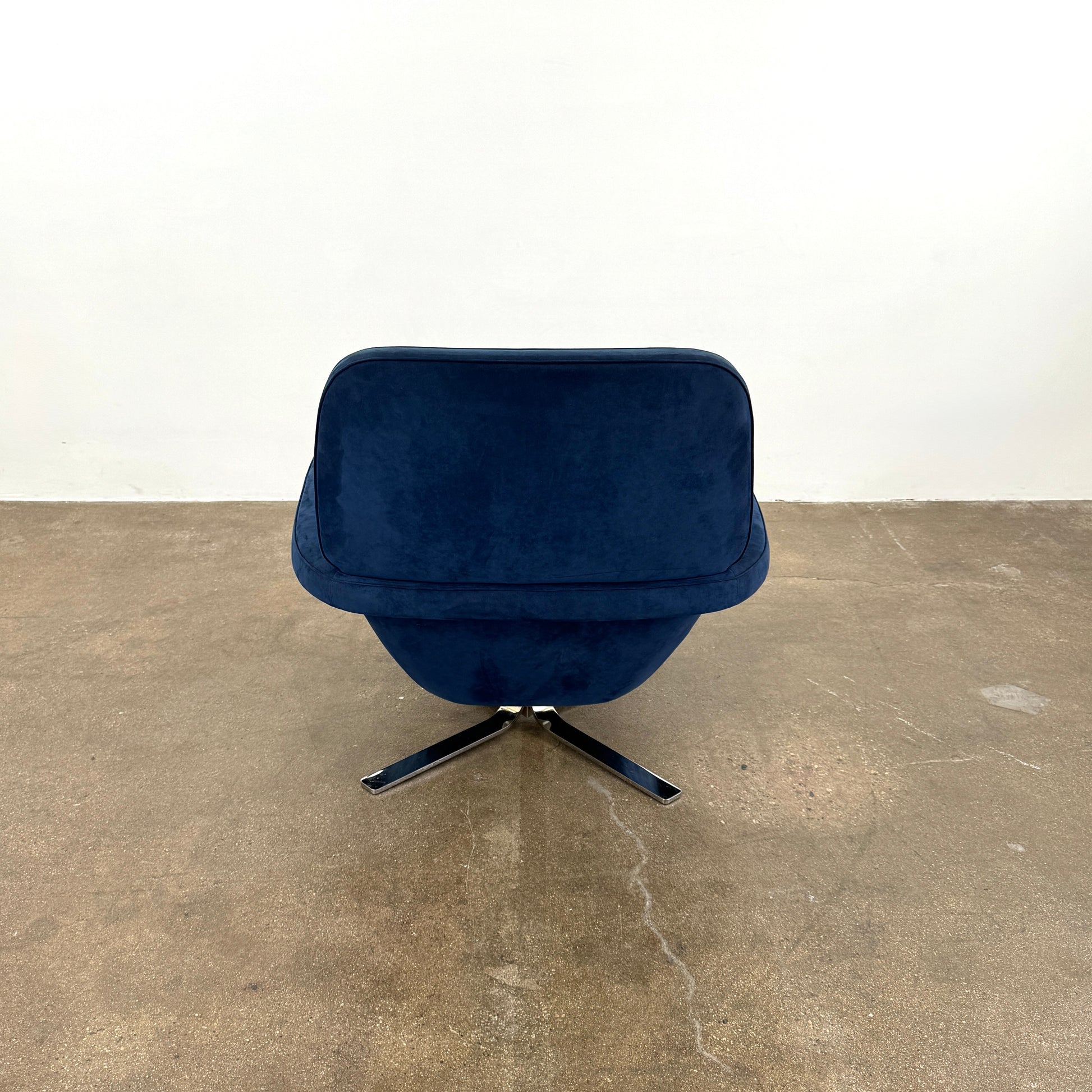 The Minotti Blake Swivel Armchair in modern navy blue, featuring a matching cushion and metal swivel base, is placed on a bare concrete floor against a plain white wall.