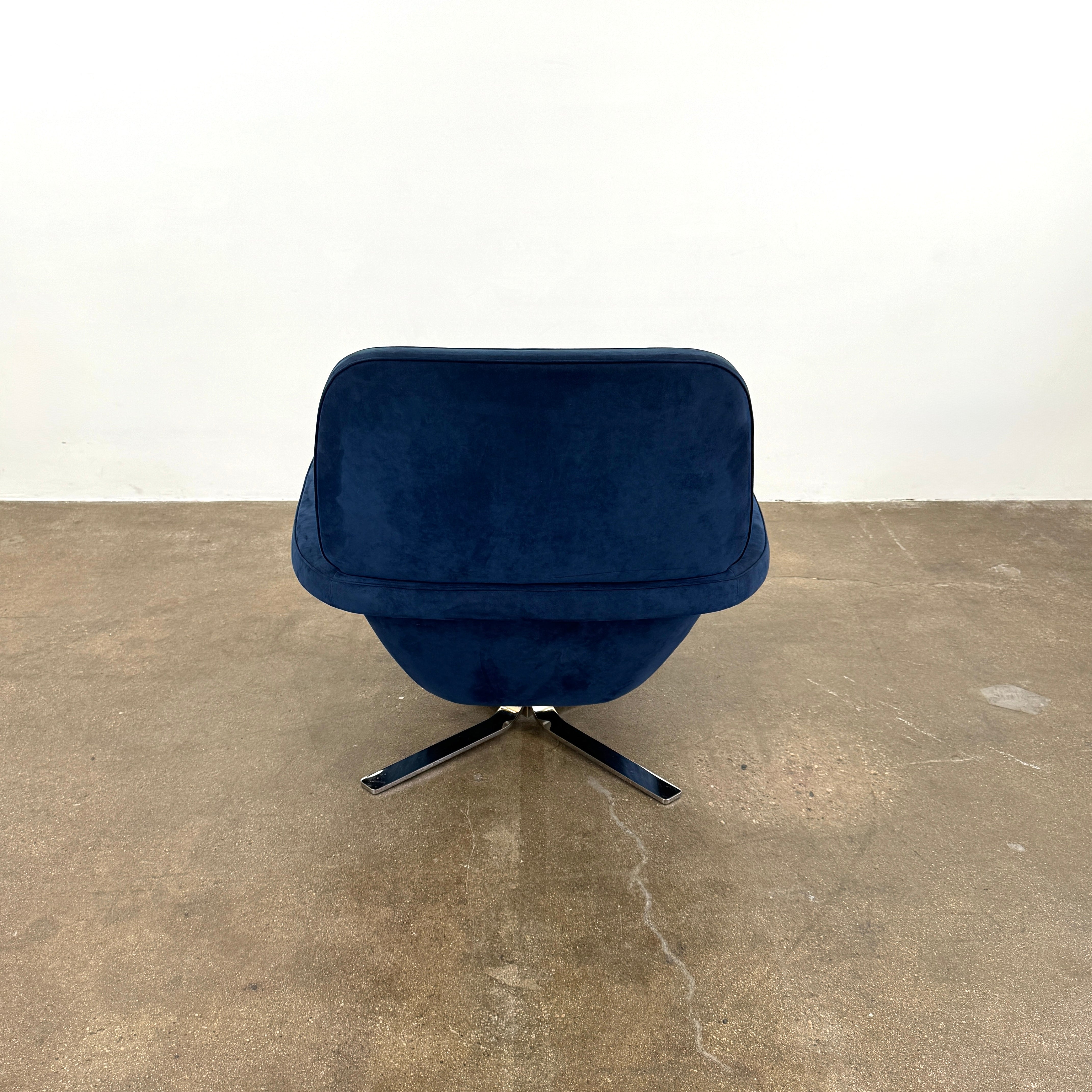 The Minotti Blake Swivel Armchair in modern navy blue, featuring a matching cushion and metal swivel base, is placed on a bare concrete floor against a plain white wall.