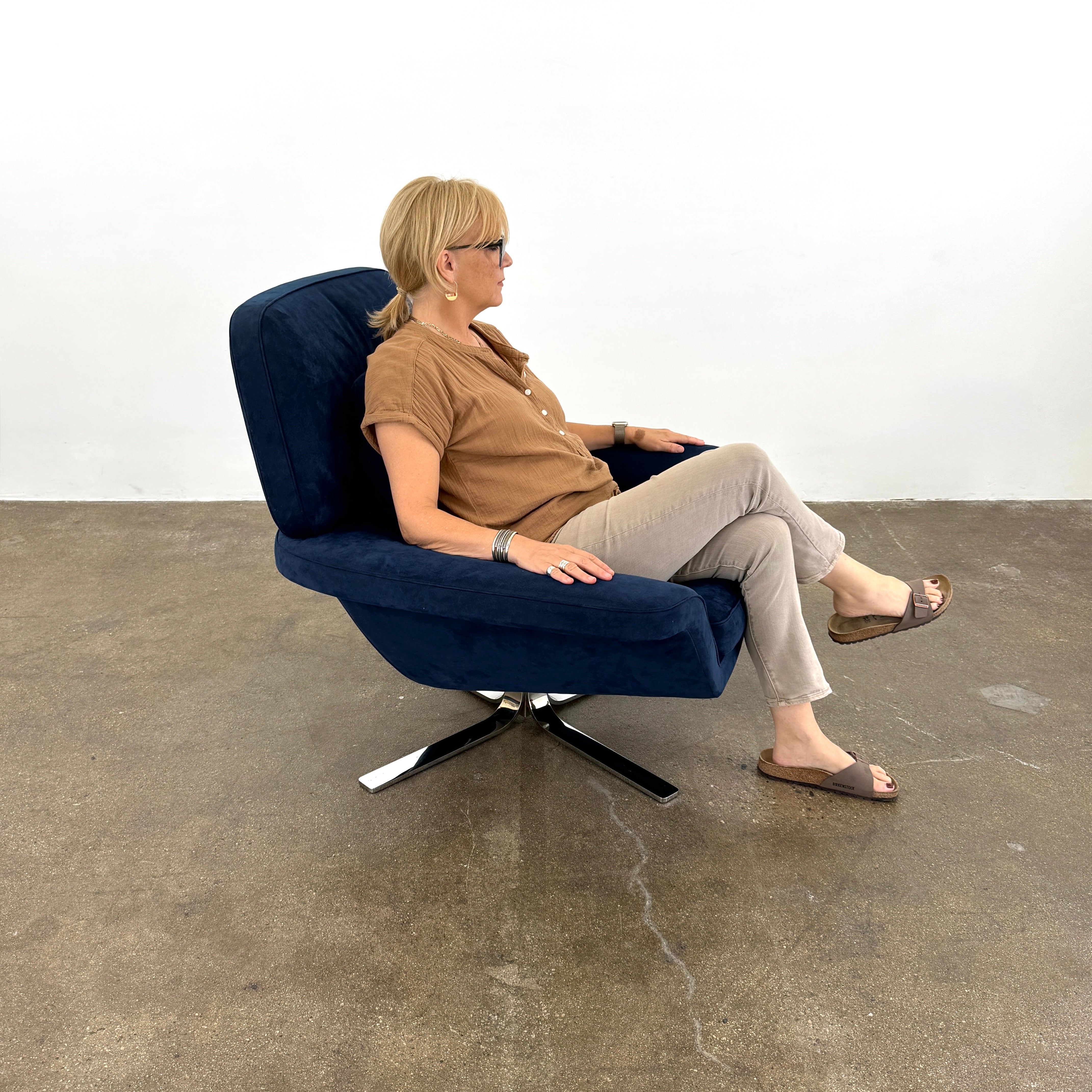 The Minotti Blake Swivel Armchair in modern navy blue, featuring a matching cushion and metal swivel base, is placed on a bare concrete floor against a plain white wall.