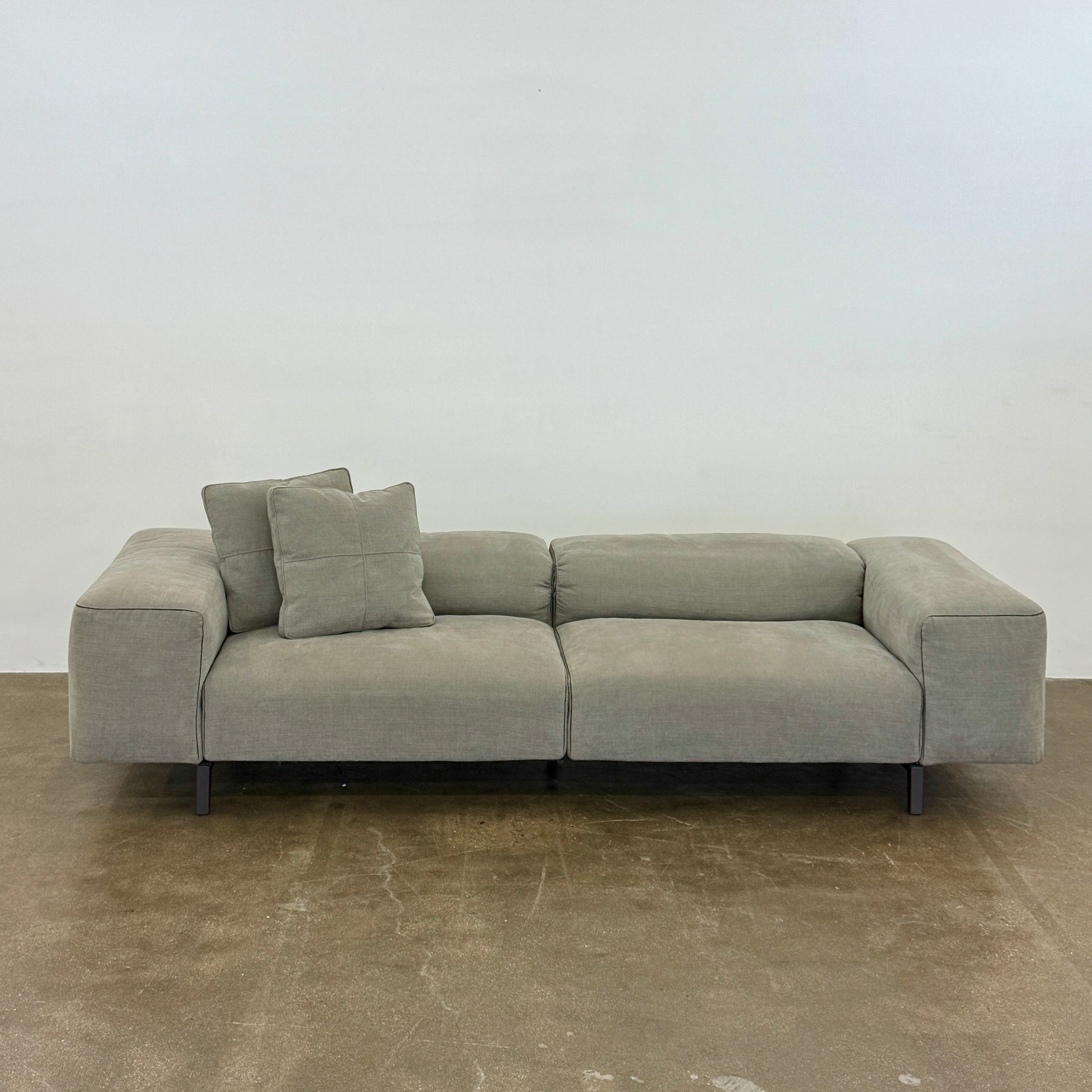A modern Cassina Scighera Sofa by Cassina, in light gray with two matching square cushions, is placed on a bare concrete floor against a plain white wall.