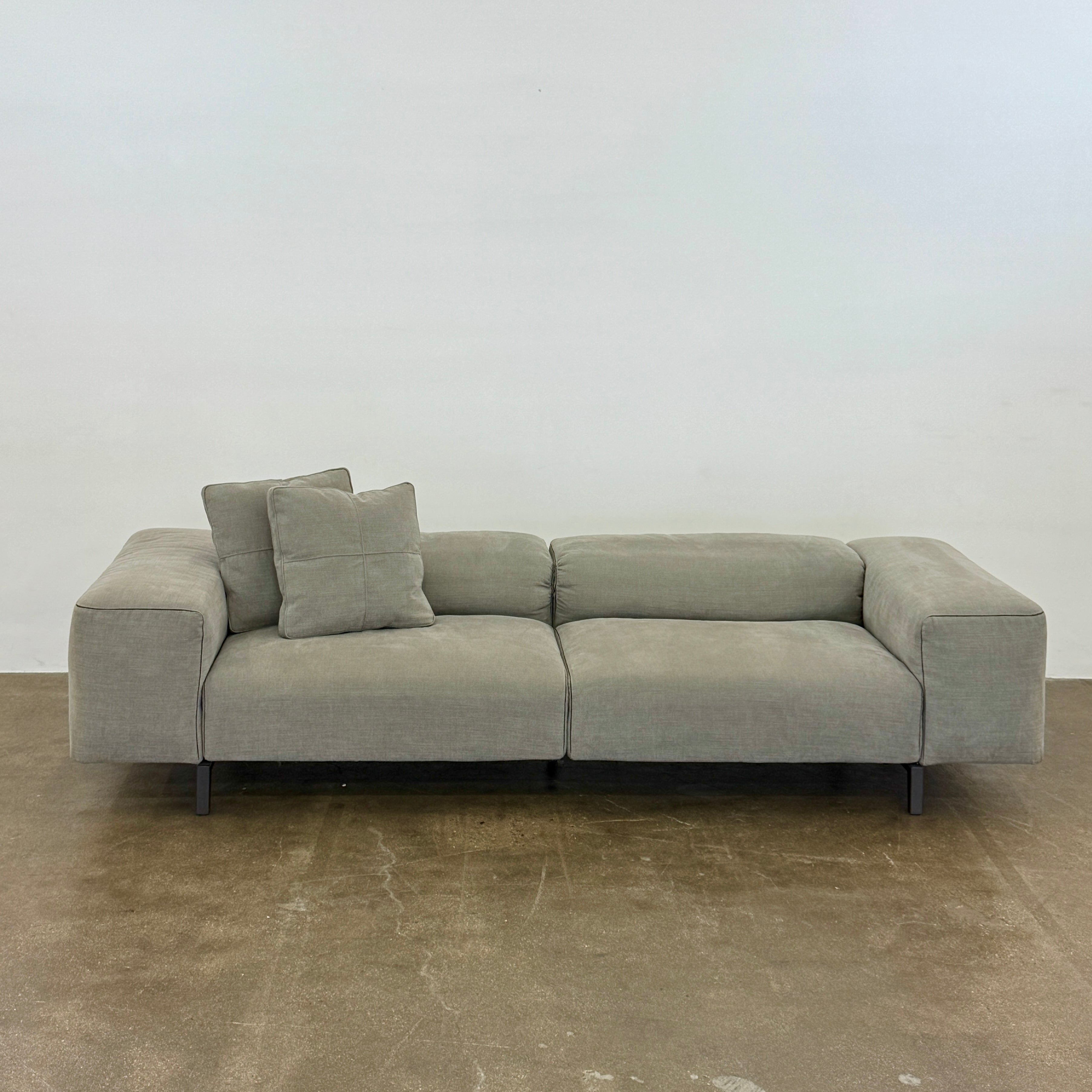 A modern Cassina Scighera Sofa by Cassina, in light gray with two matching square cushions, is placed on a bare concrete floor against a plain white wall.