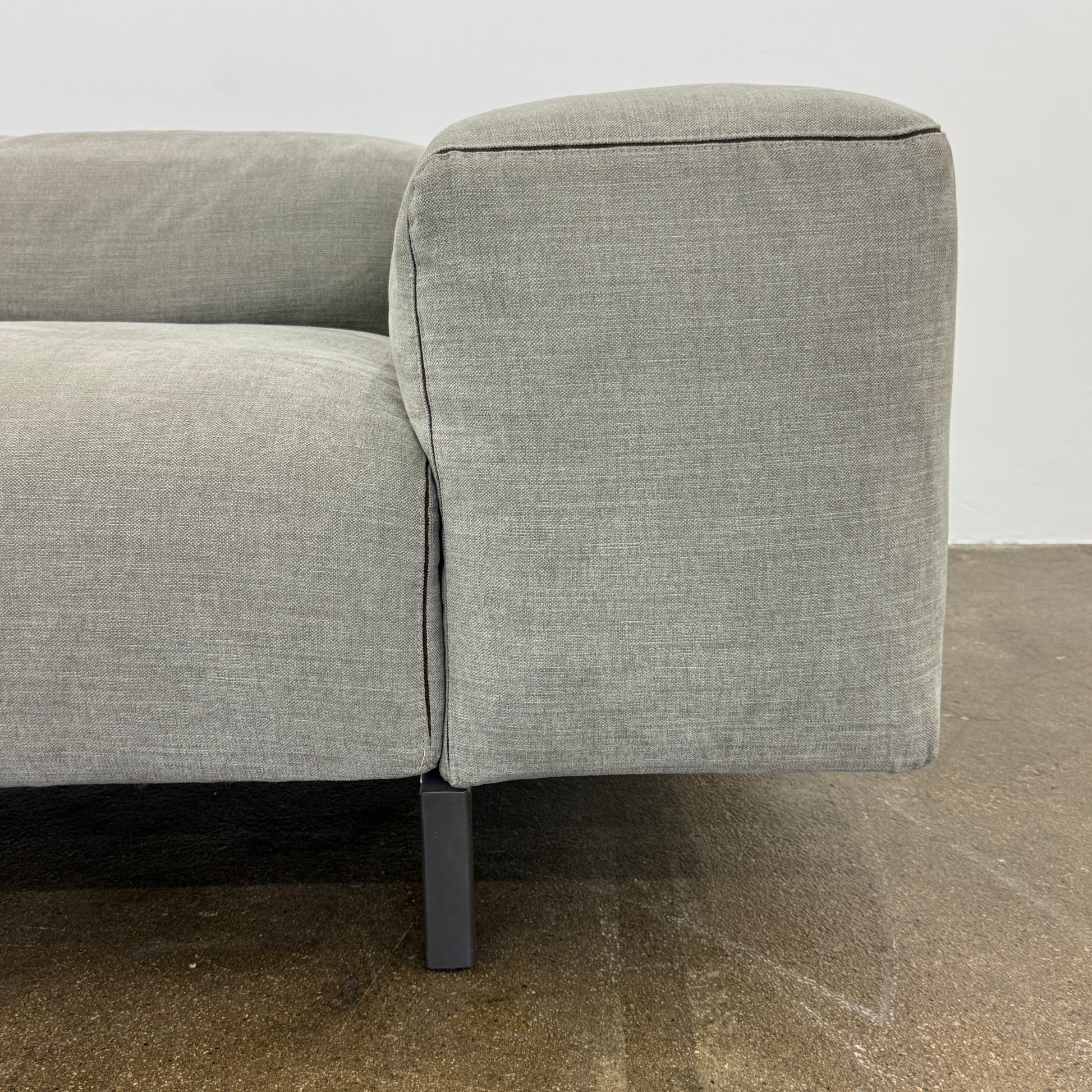 A modern Cassina Scighera Sofa by Cassina, in light gray with two matching square cushions, is placed on a bare concrete floor against a plain white wall.
