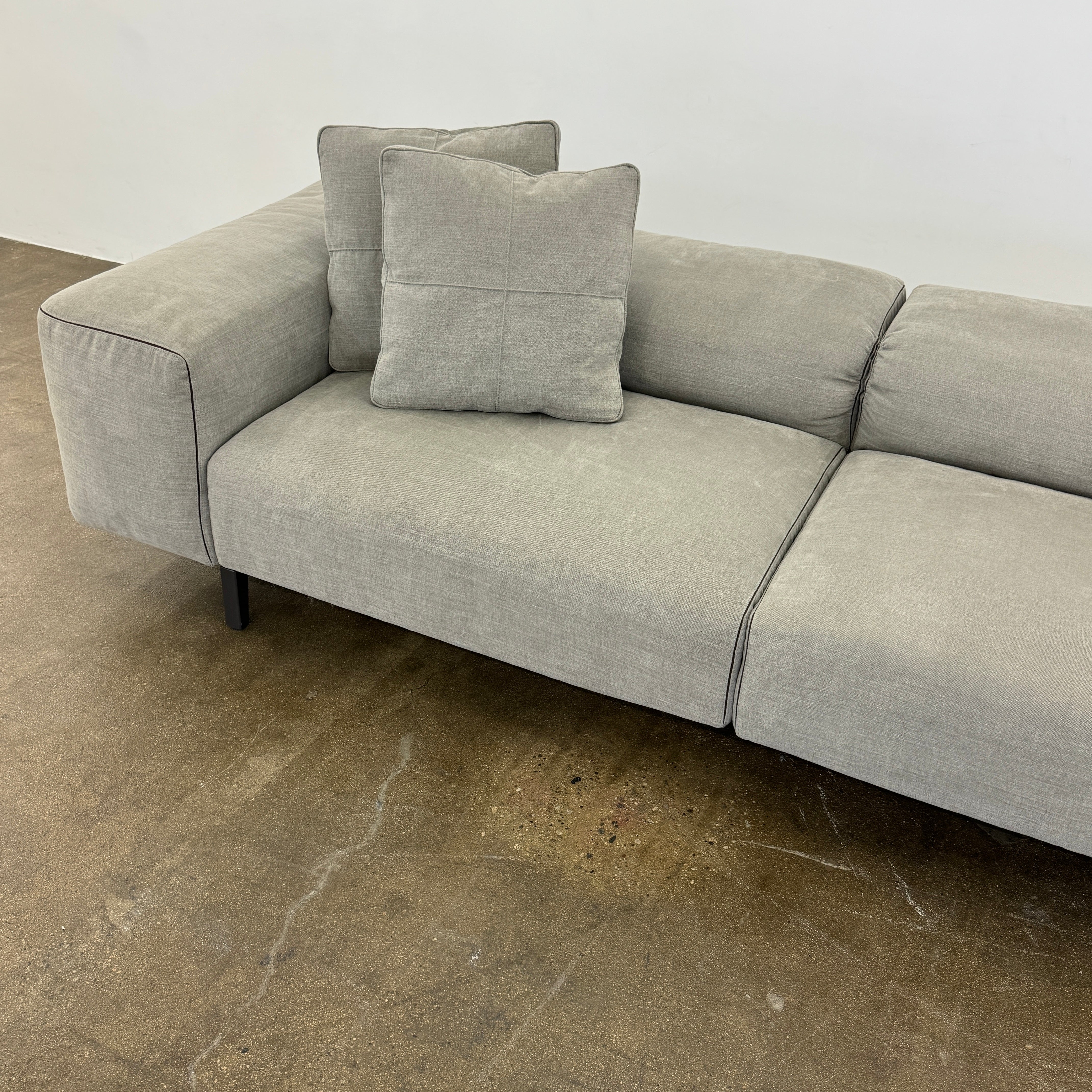 A modern Cassina Scighera Sofa by Cassina, in light gray with two matching square cushions, is placed on a bare concrete floor against a plain white wall.