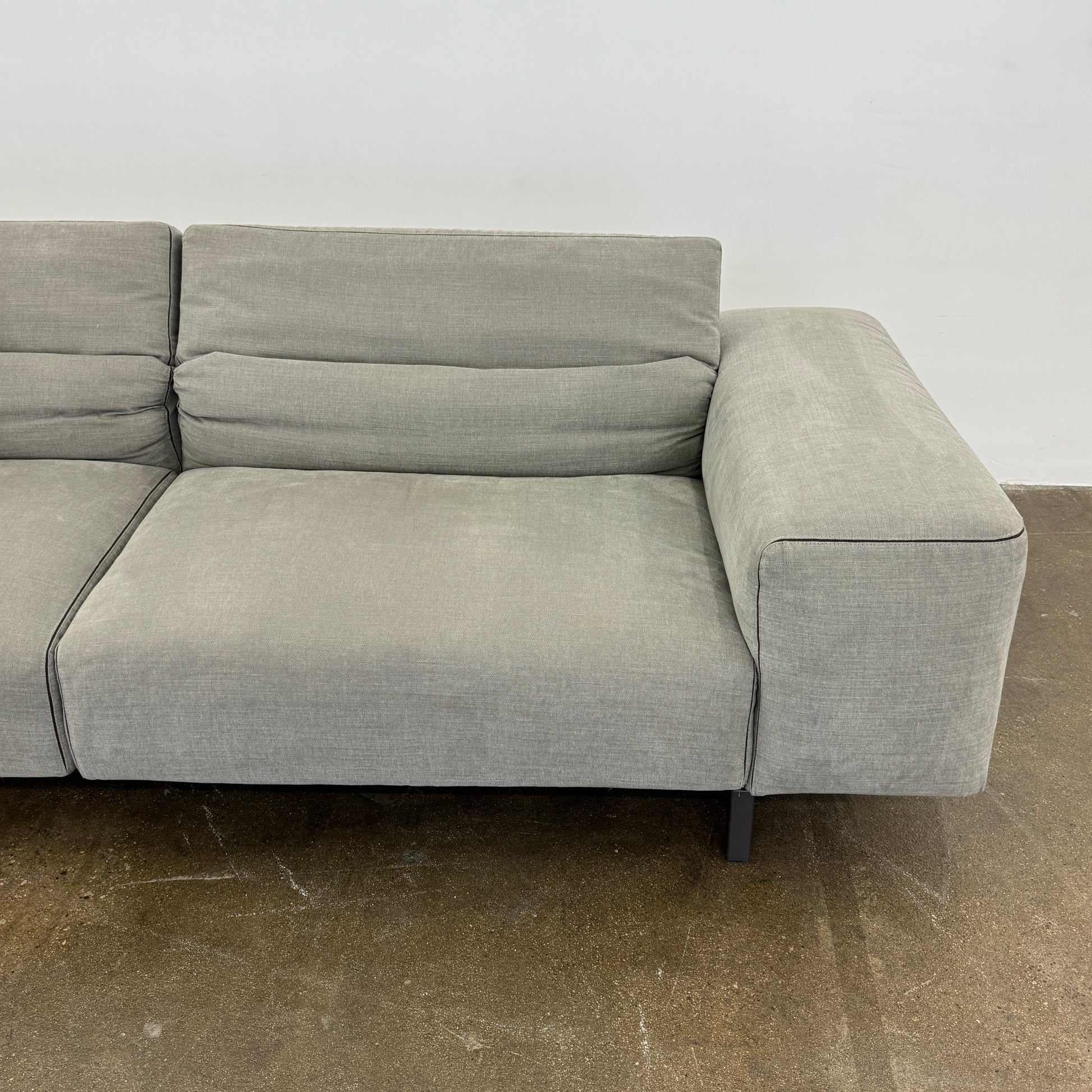 A modern Cassina Scighera Sofa by Cassina, in light gray with two matching square cushions, is placed on a bare concrete floor against a plain white wall.
