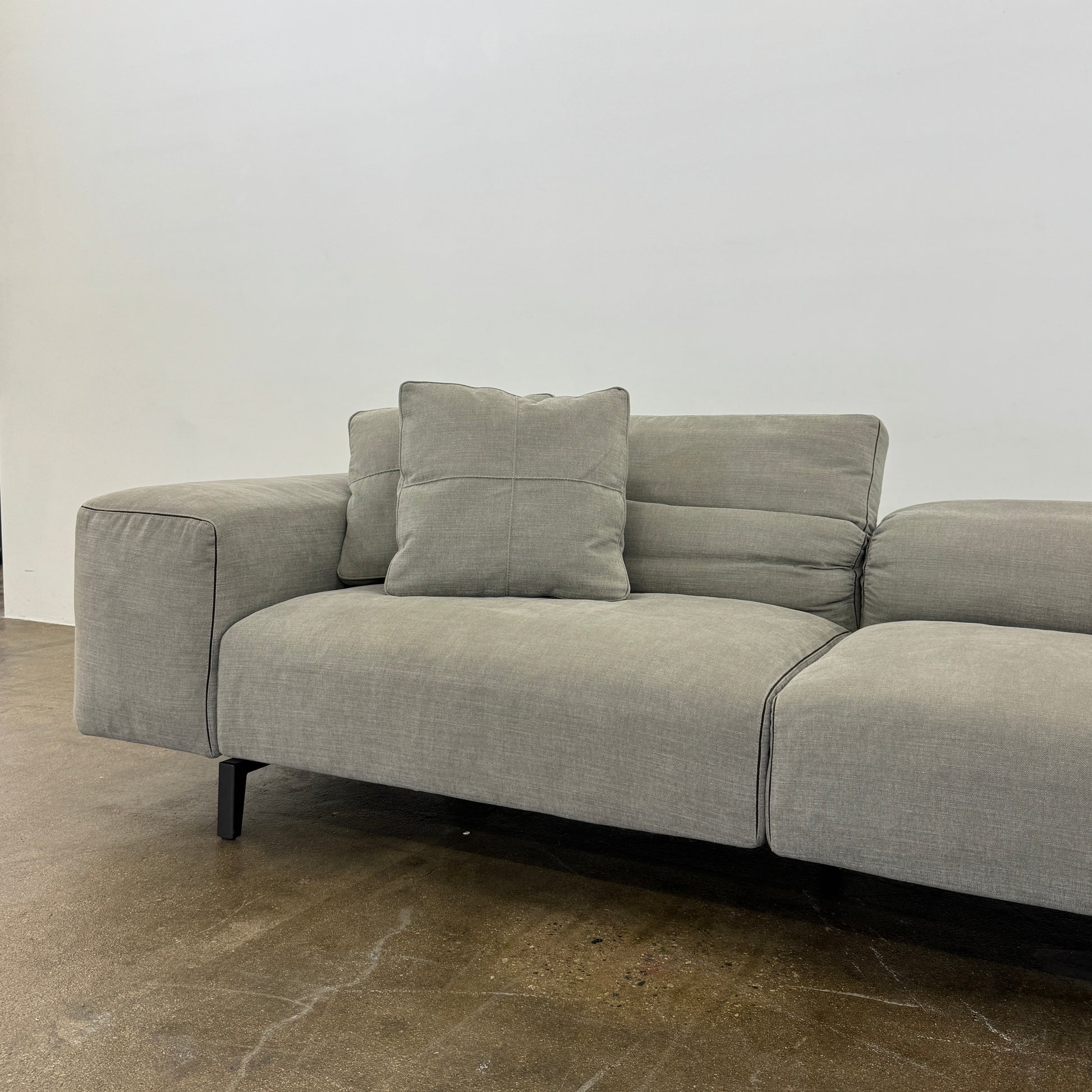 A modern Cassina Scighera Sofa by Cassina, in light gray with two matching square cushions, is placed on a bare concrete floor against a plain white wall.